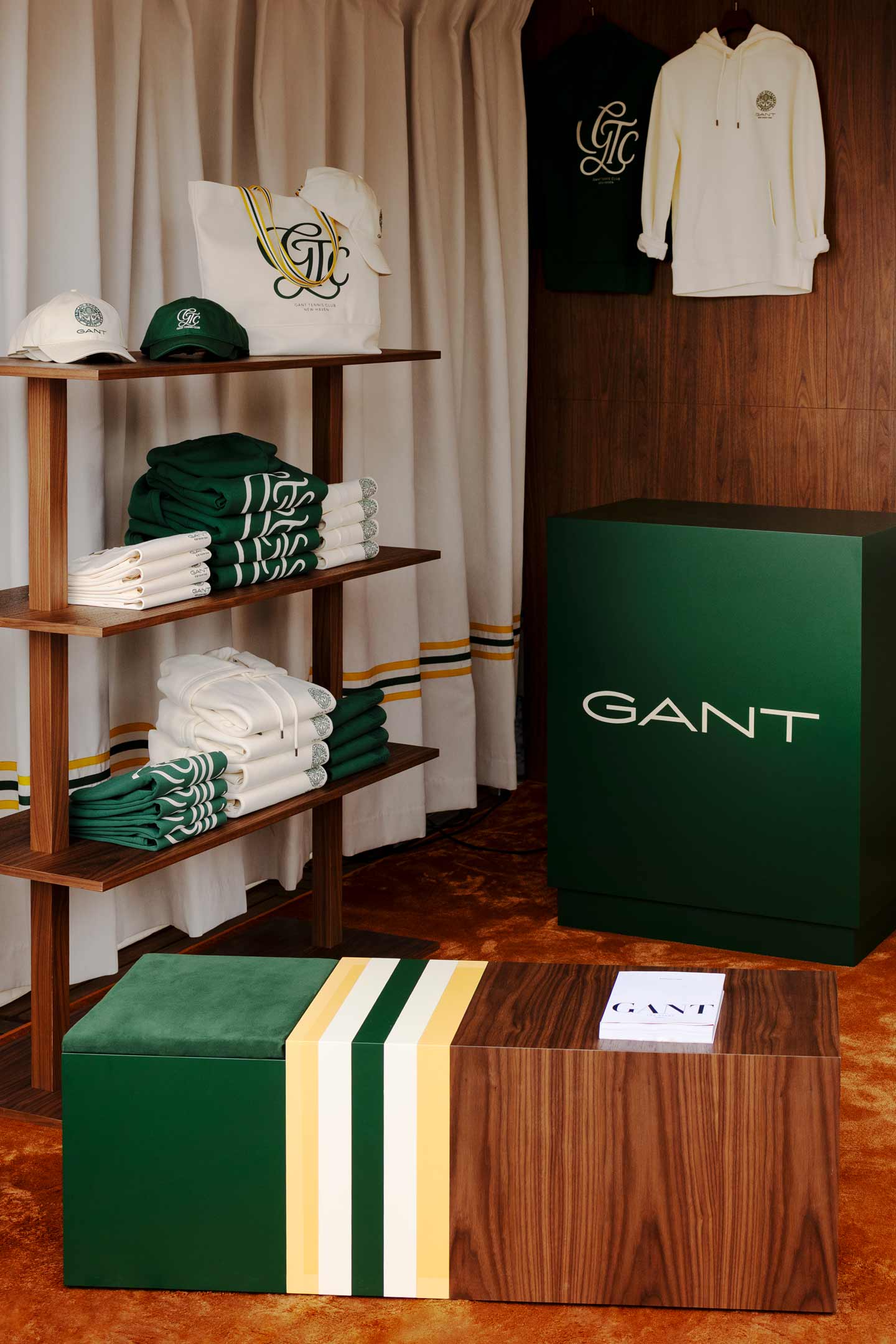 GANT American Sports Center pop-up display featuring folded sweaters, polo shirts, hoodies, caps, tennis-inspired apparel, and mannequins against a wooden backdrop with the GANT tennis club emblem.