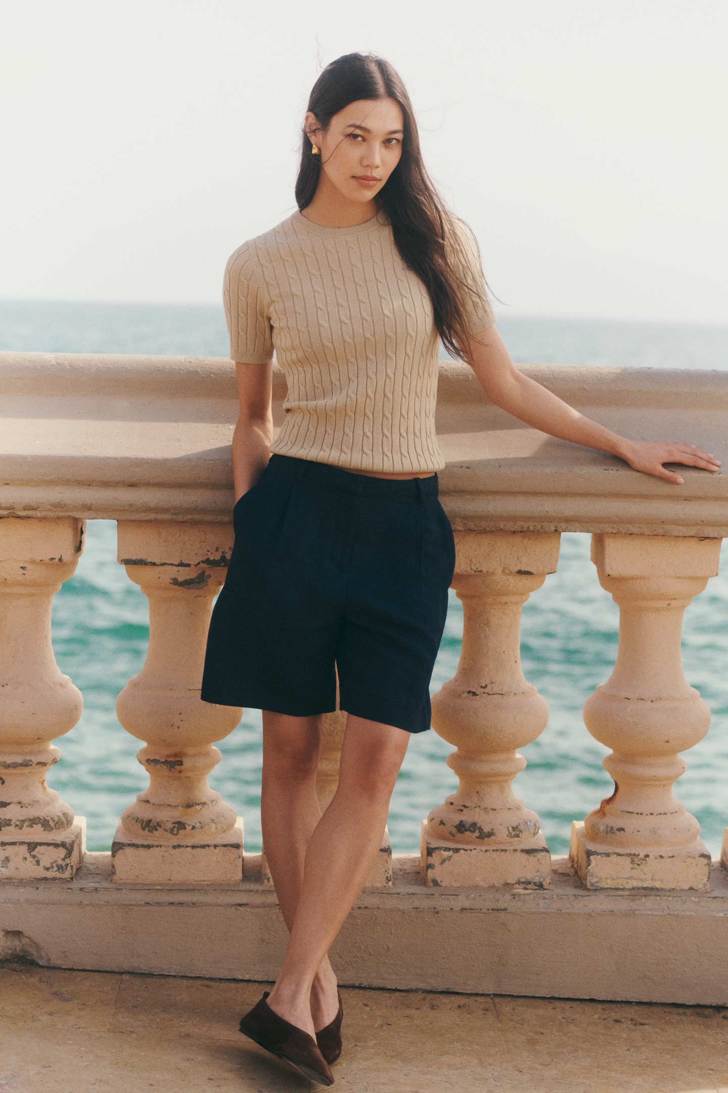 Woman wearing a Gant beige short-sleeve cable-knit top and dark shorts standing by a seaside railing.