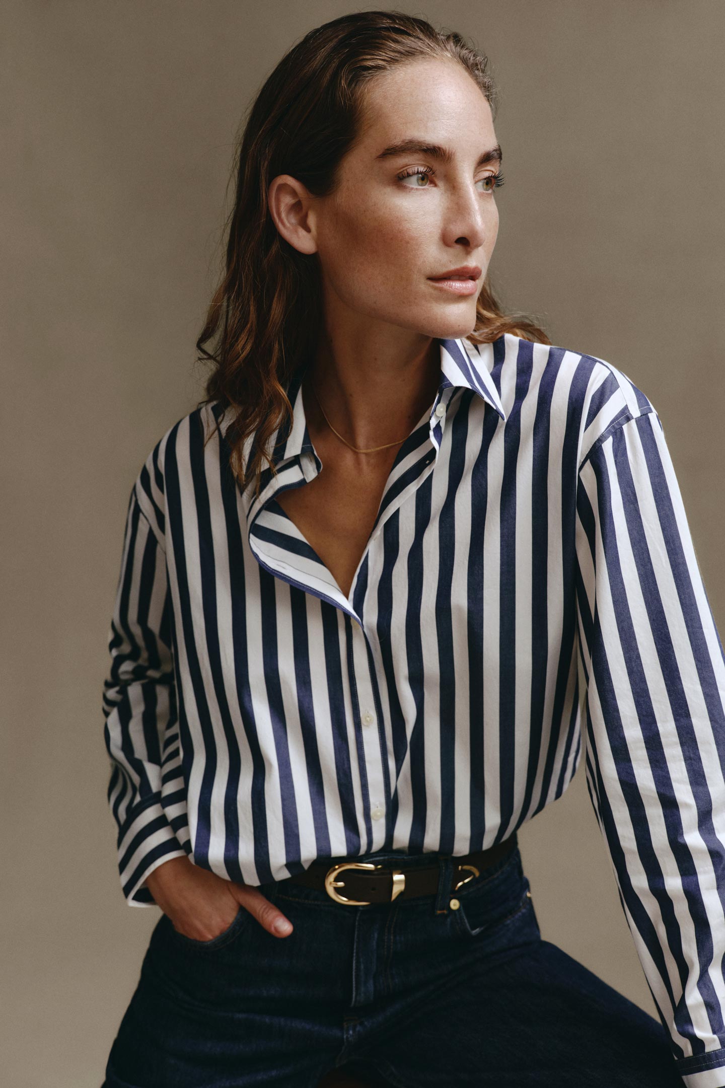 Model styled in a Gant striped button-down shirt tucked into dark-wash jeans, seated in a studio setting.