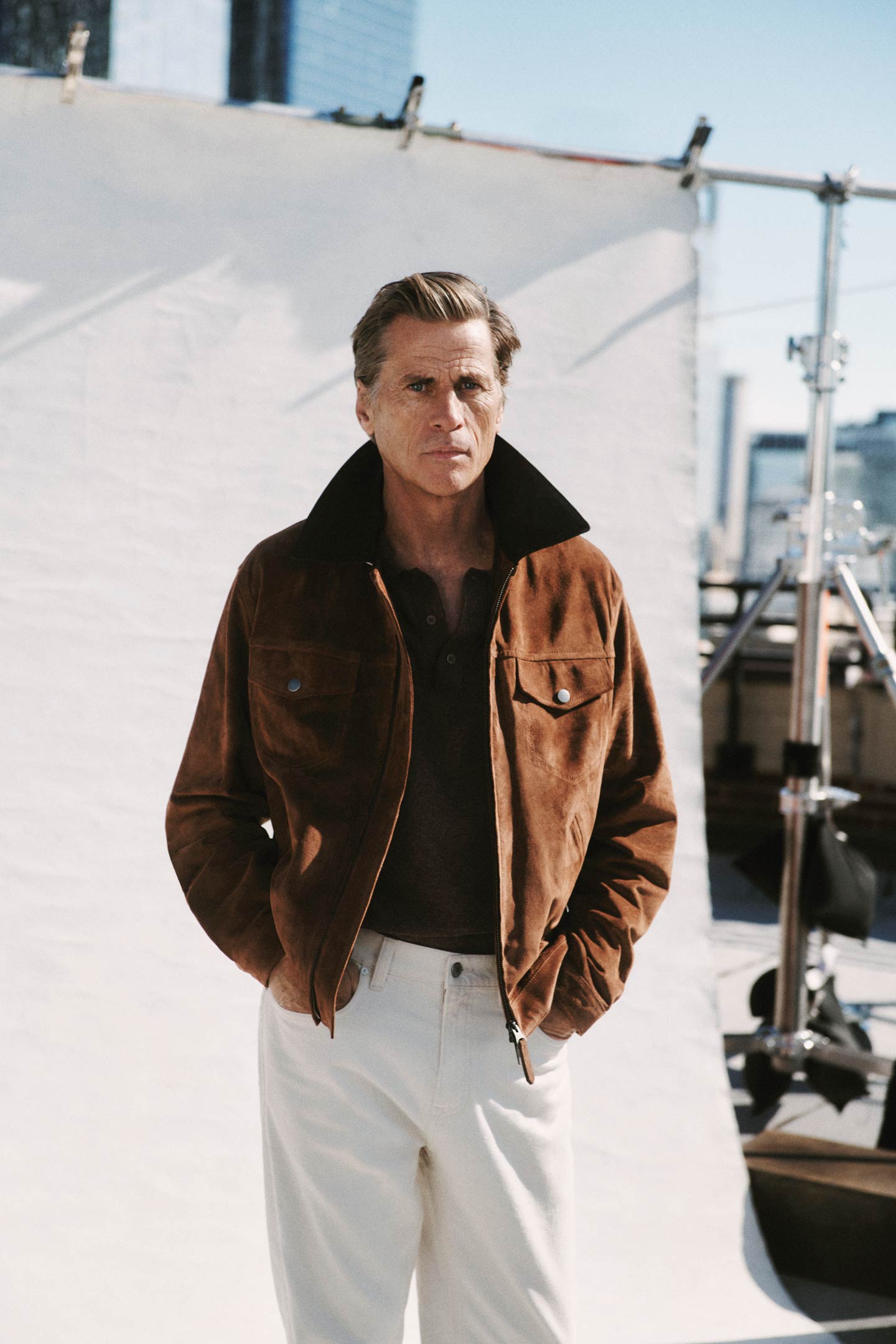 Person wearing a brown suede Gant jacket over a dark shirt paired with white trousers on a rooftop.