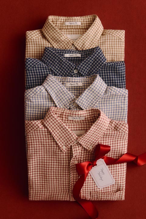 Stack of folded checked button-down shirts in cream, navy, blue, and red tones, styled with a gift ribbon.