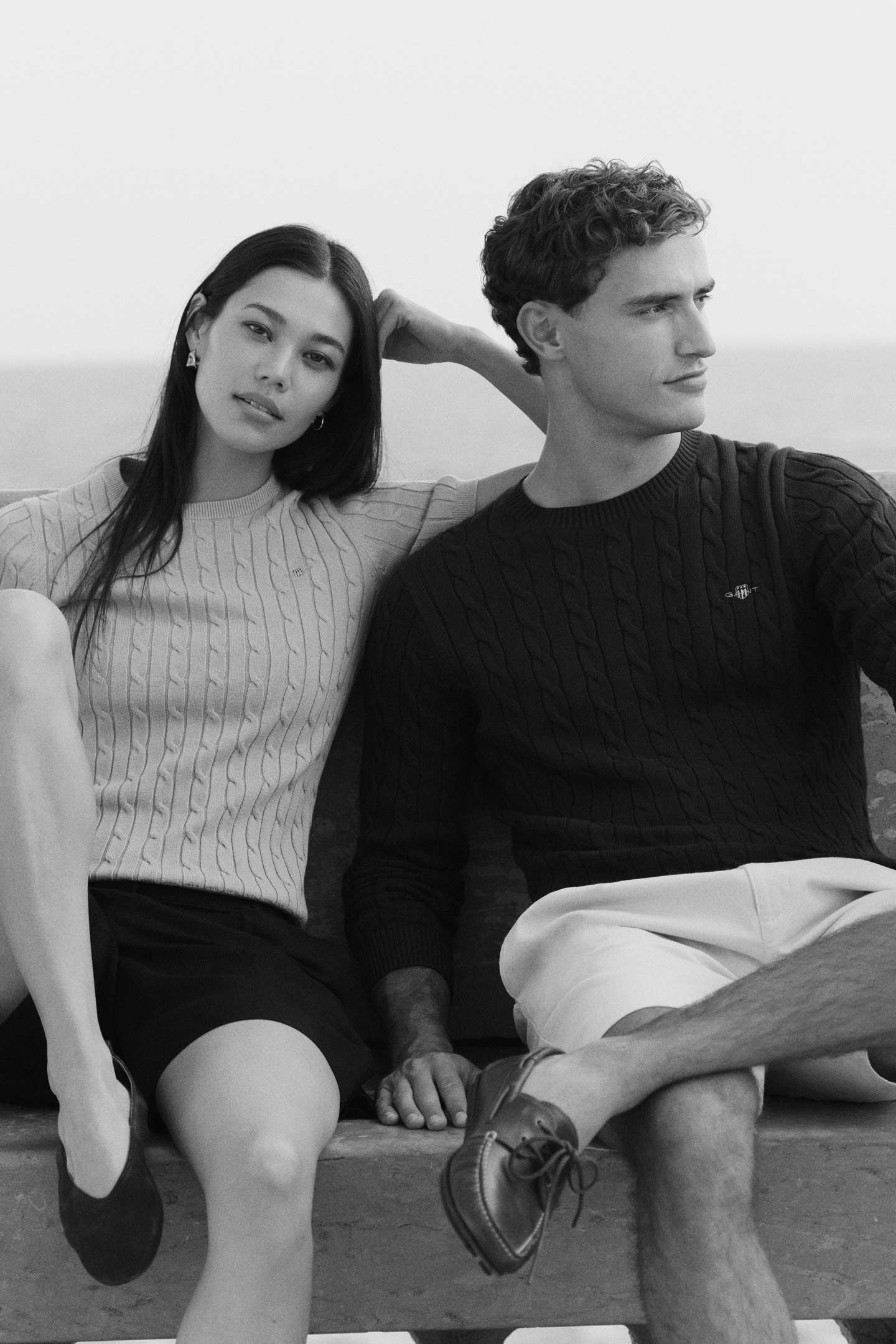 Two people wearing Gant cable-knit sweaters and shorts seated together on a stone bench by the water.