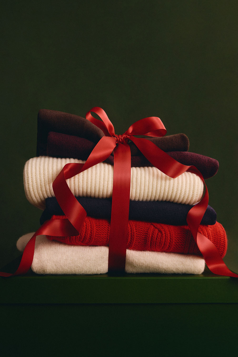 Stack of folded knitwear in cream, navy, red and burgundy, wrapped with a red gift ribbon.