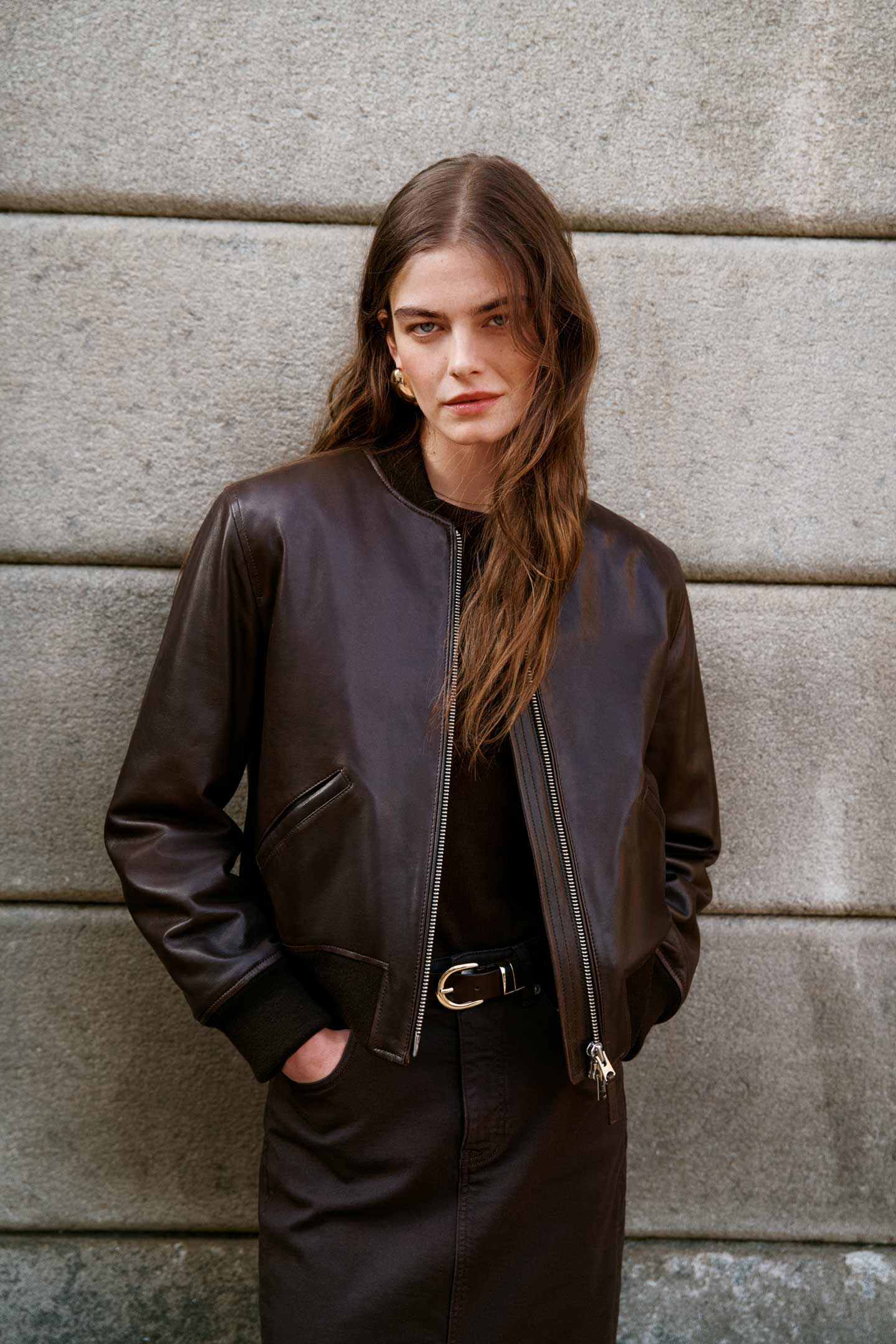 Model styled in a GANT leather jacket over layered outfit, standing against a stone wall outdoors.