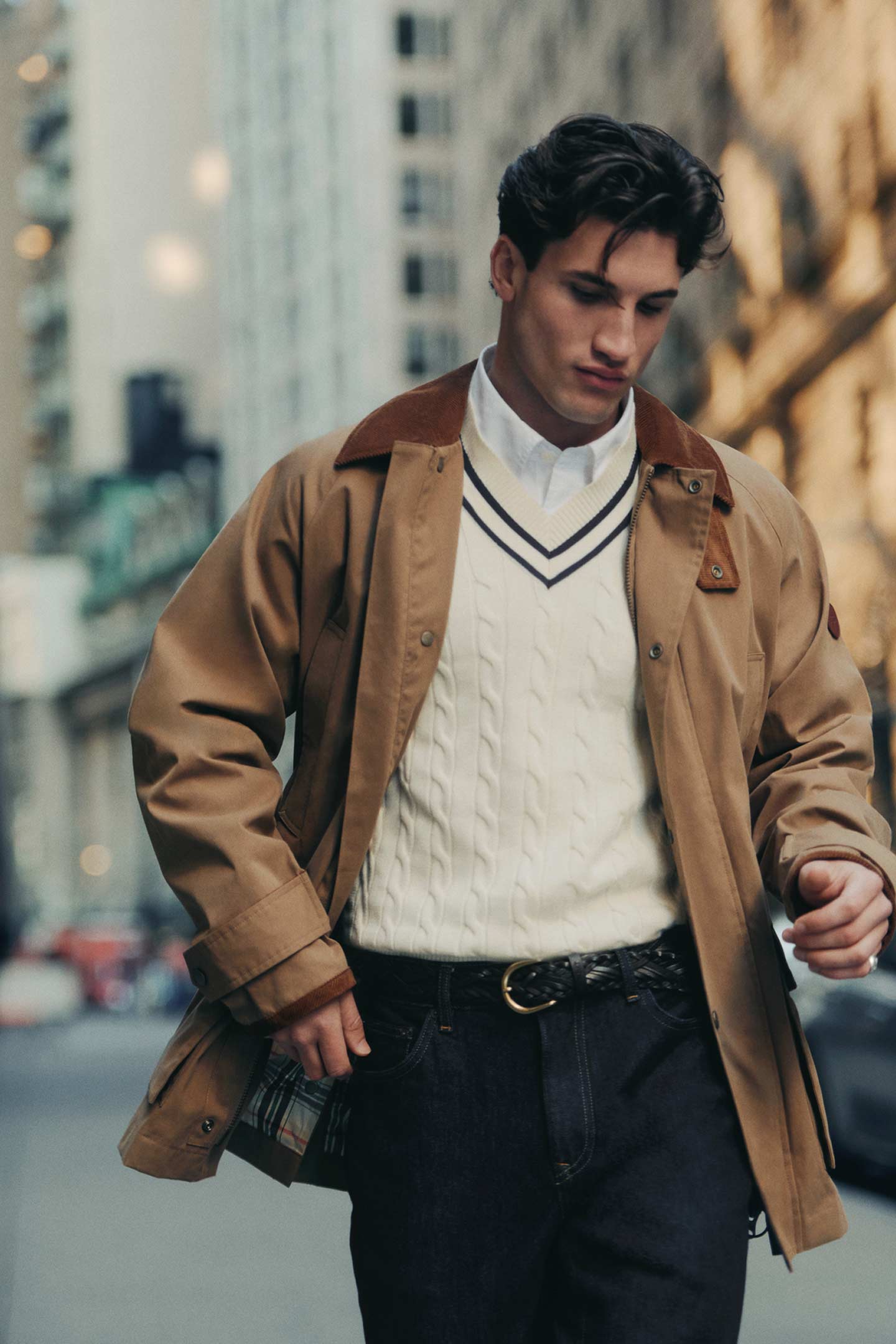 Personne portant un pull en tricot torsadé crème Gant sous une veste beige en se promenant dans un cadre urbain.