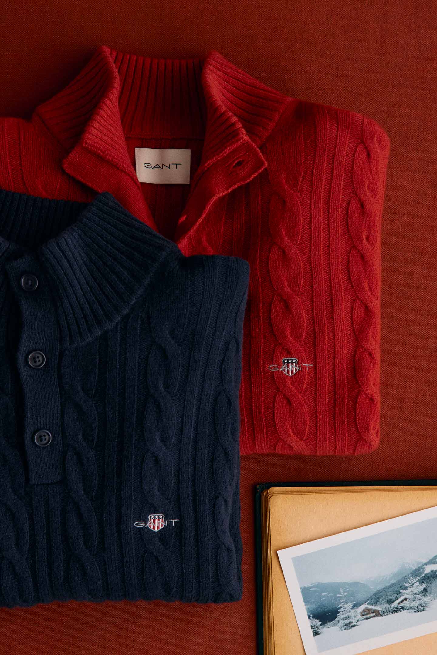 Folded red cable-knit sweater layered with a navy knit polo shirt, both featuring embroidered Gant shield logos.