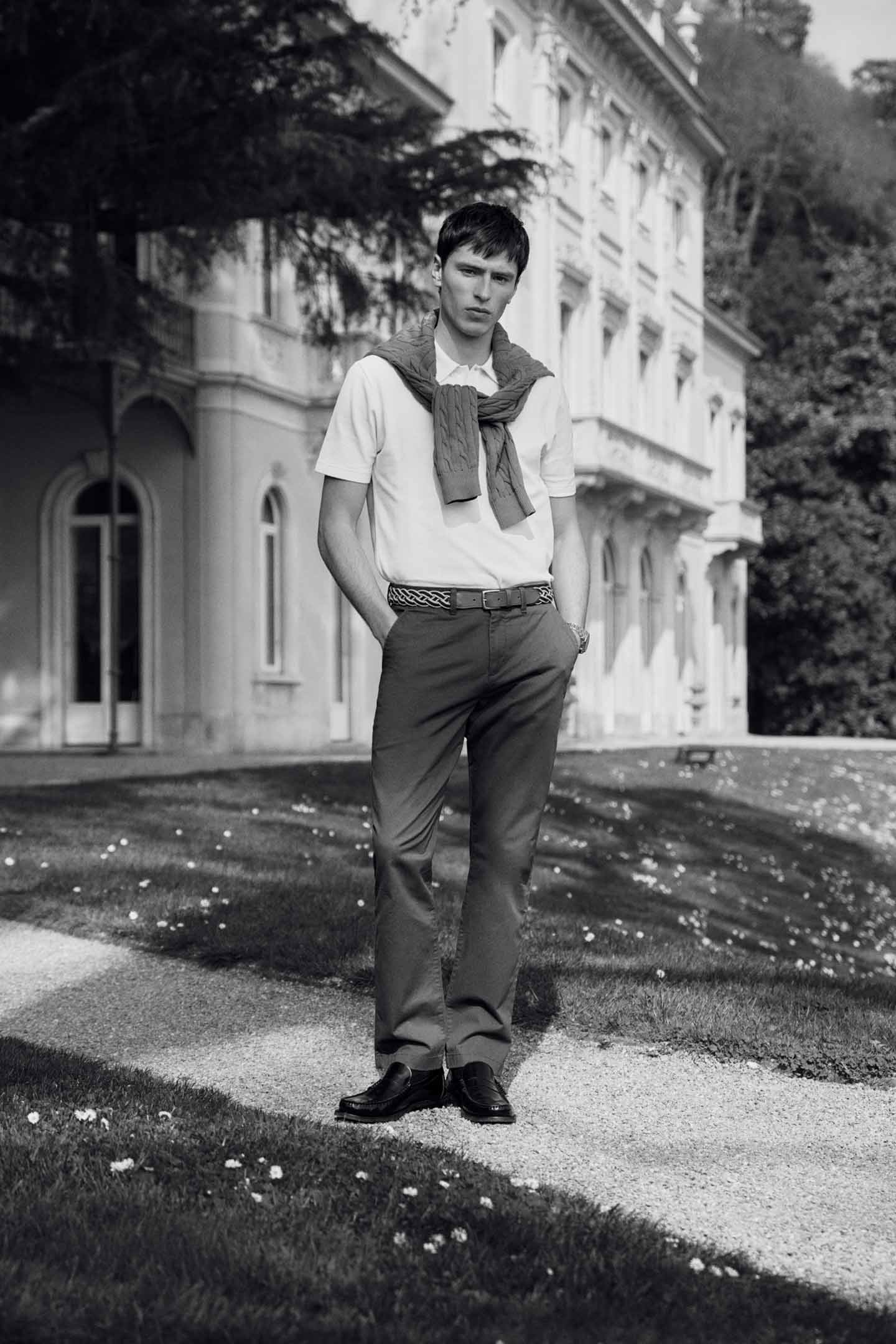 Black and white image of a model styled in a GANT polo shirt and tailored trousers standing outdoors near a classic building.