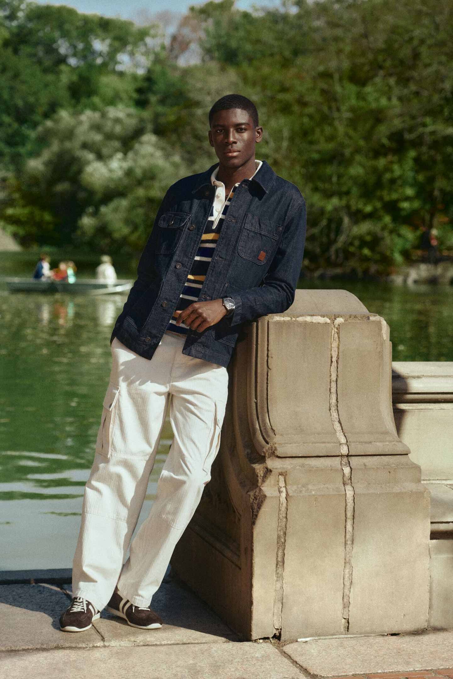 Model wearing a dark denim jacket layered over a striped shirt, paired with white cargo pants and brown sneakers, standing casually against a stone railing beside a lakeside park setting.