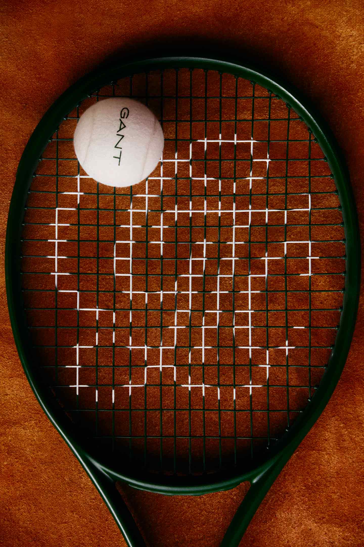 Tennis racket with GANT‑branded tennis ball on a clay‑colored surface.
