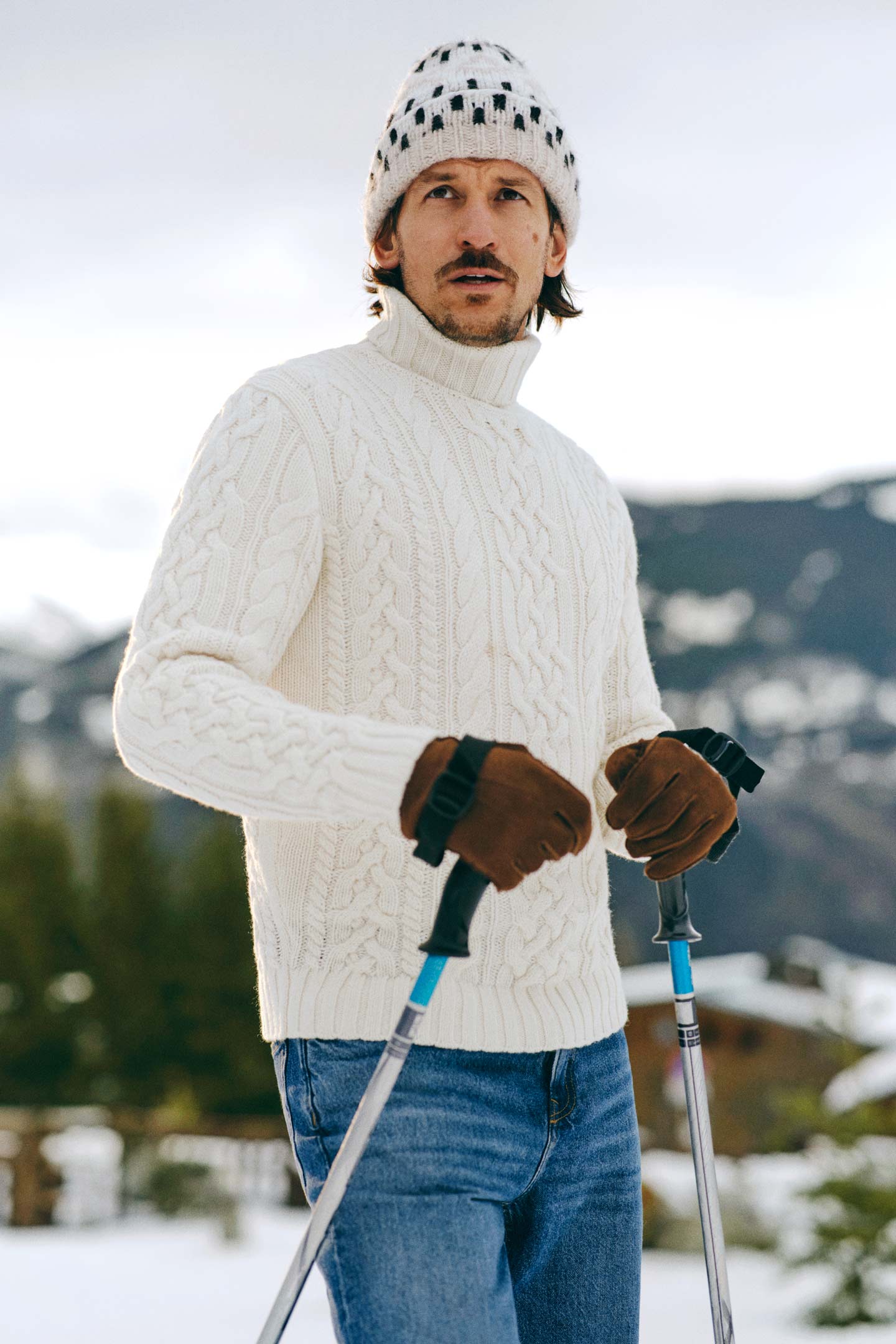 Model styled in a Gant cable-knit roll-neck sweater paired with jeans, gloves and a knitted beanie in the snow.