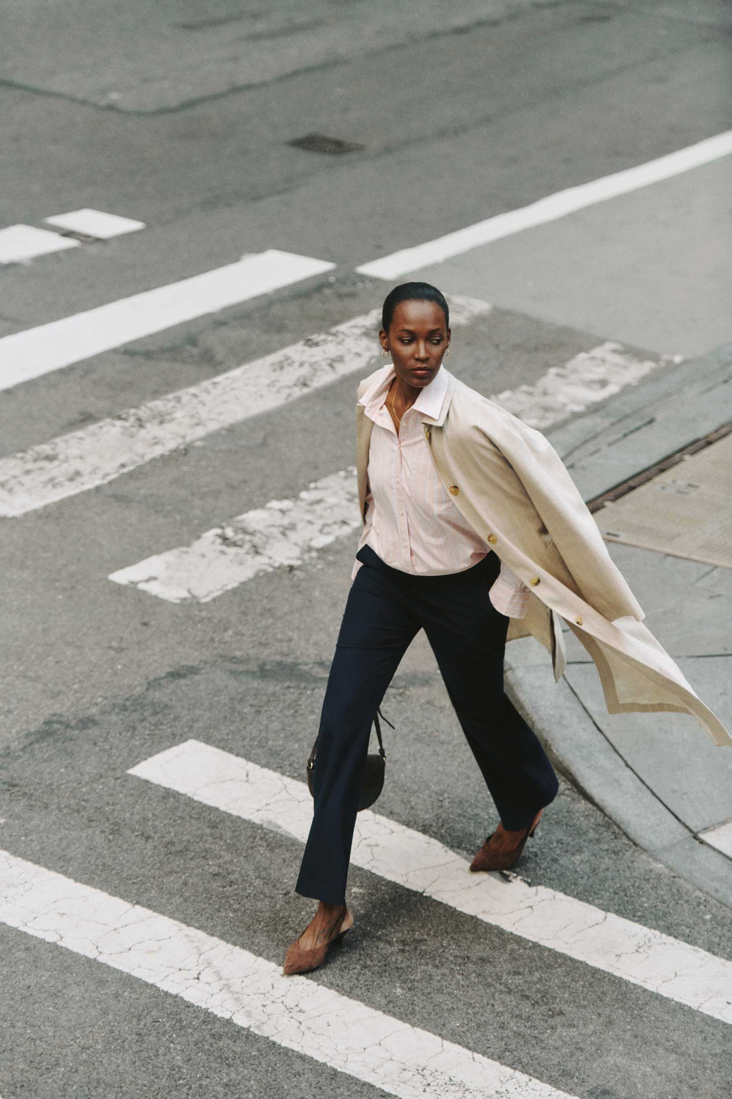 Woman wearing a Gant light coat and shirt with navy trousers while walking across a city street.