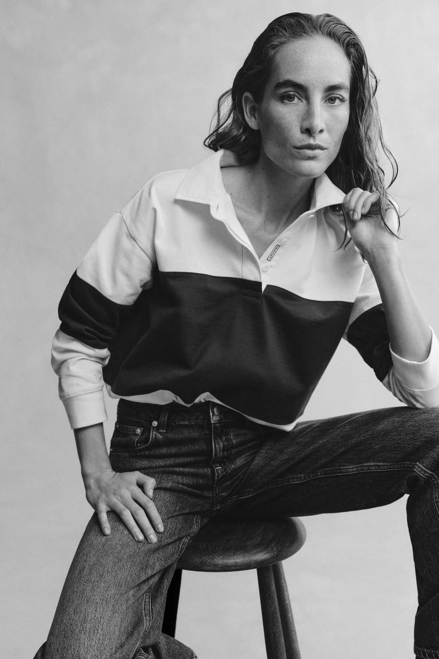 Black and white image of a model styled in a GANT color-blocked rugger layered over a shirt with jeans sitting on a stool.