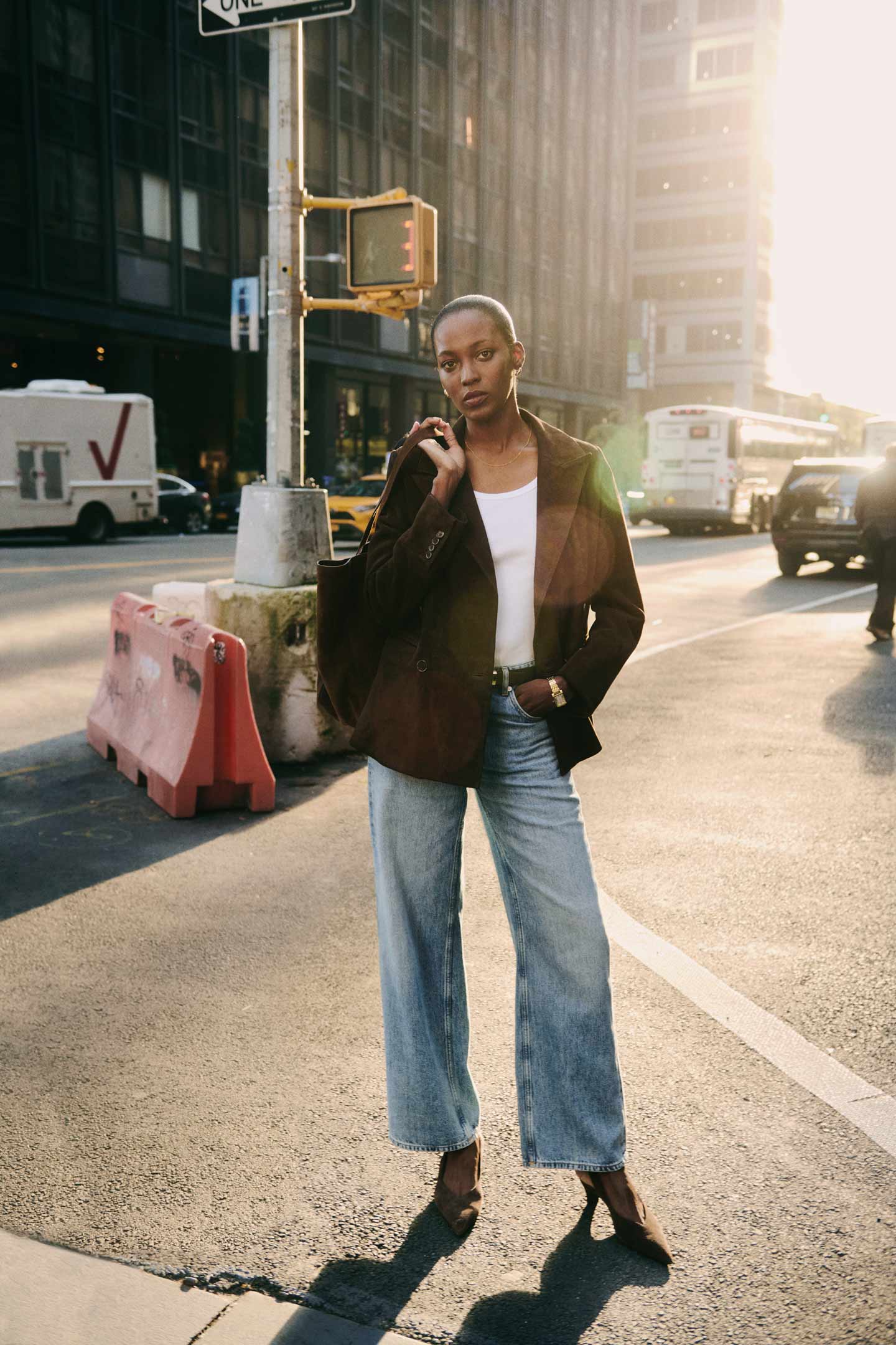 Woman wearing a brown Gant blazer, white top and wide-leg jeans standing at a city crosswalk.