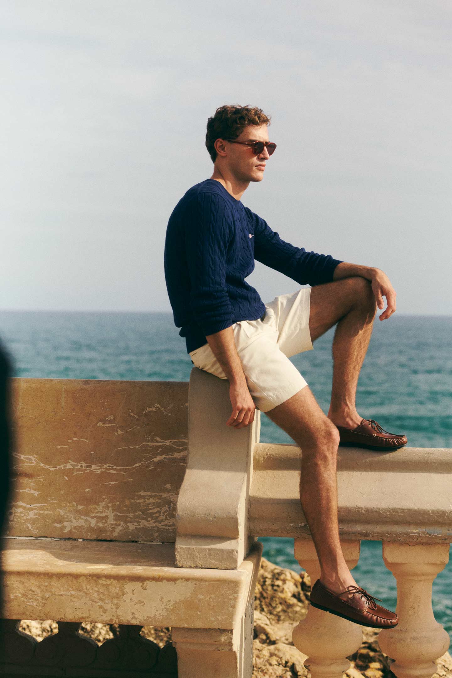 Man wearing a Gant navy sweater and light shorts while seated on a stone ledge near the sea.