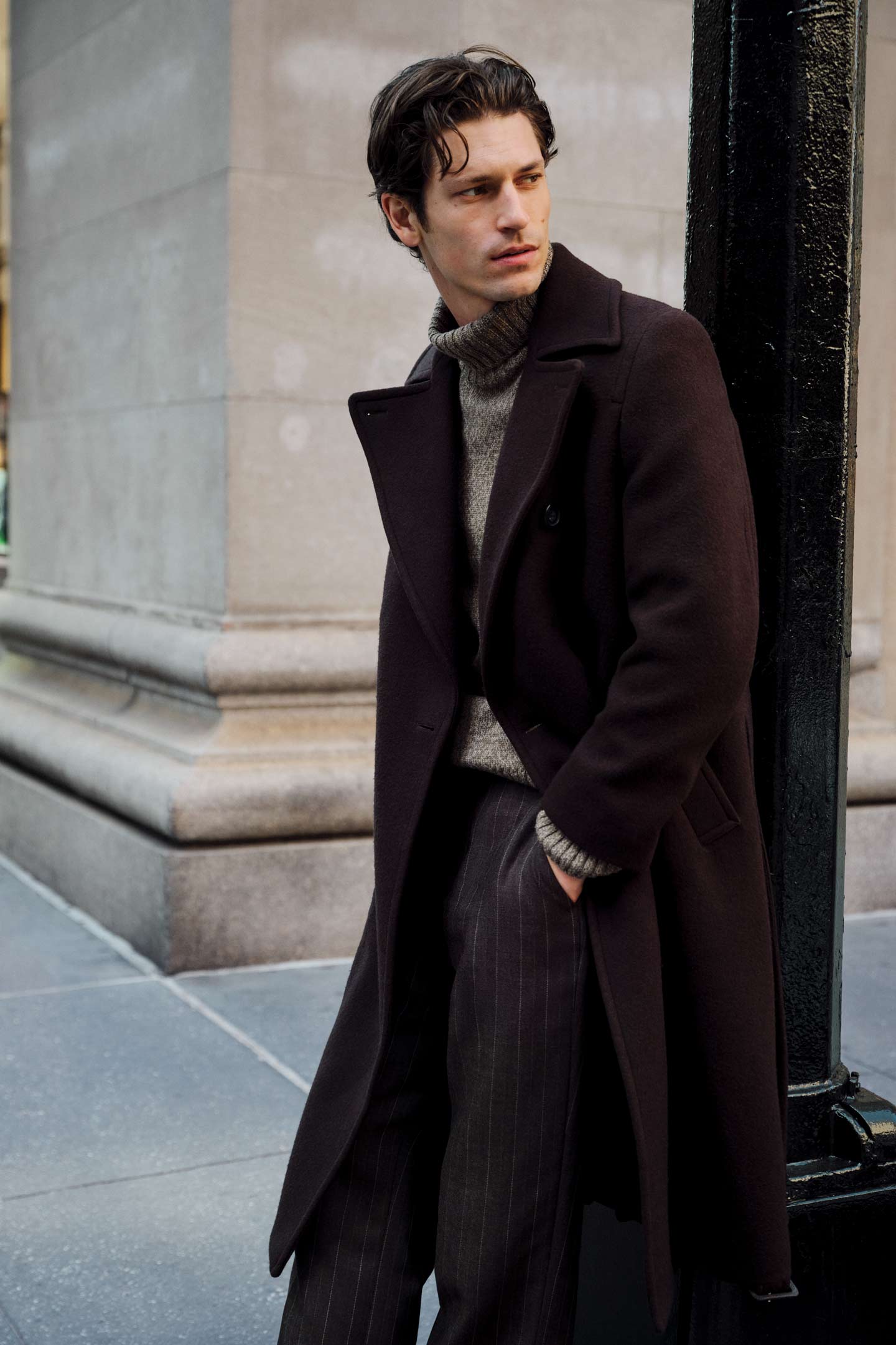 Model styled in a Gant wool overcoat worn with a roll-neck sweater and pinstripe pants, leaning against a building.