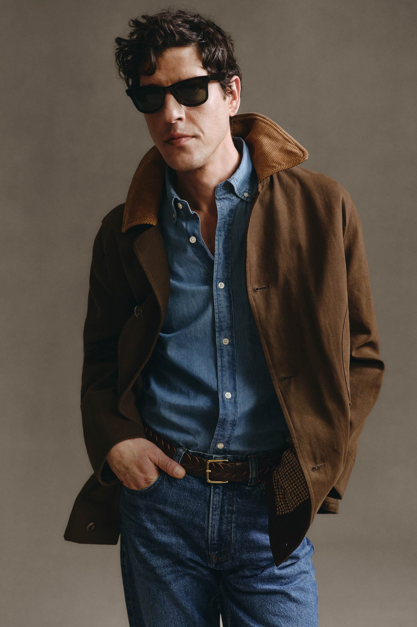 Model styled in a Gant jacket layered over a chambray shirt and jeans, styled with sunglasses in a studio setting.