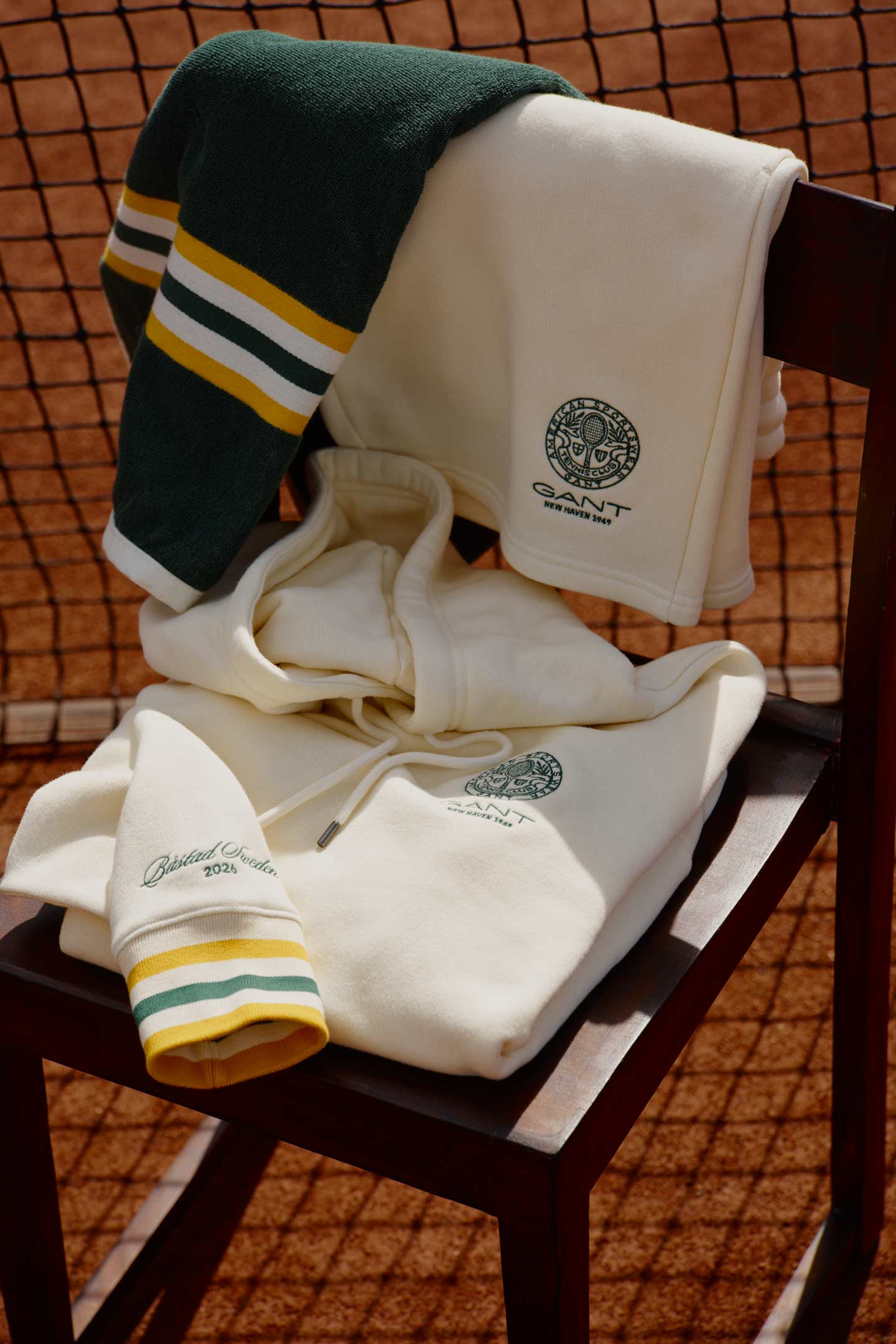 Gant tennis clothing and knitwear displayed on a chair with a tote bag and tennis balls beside a court net outdoors.