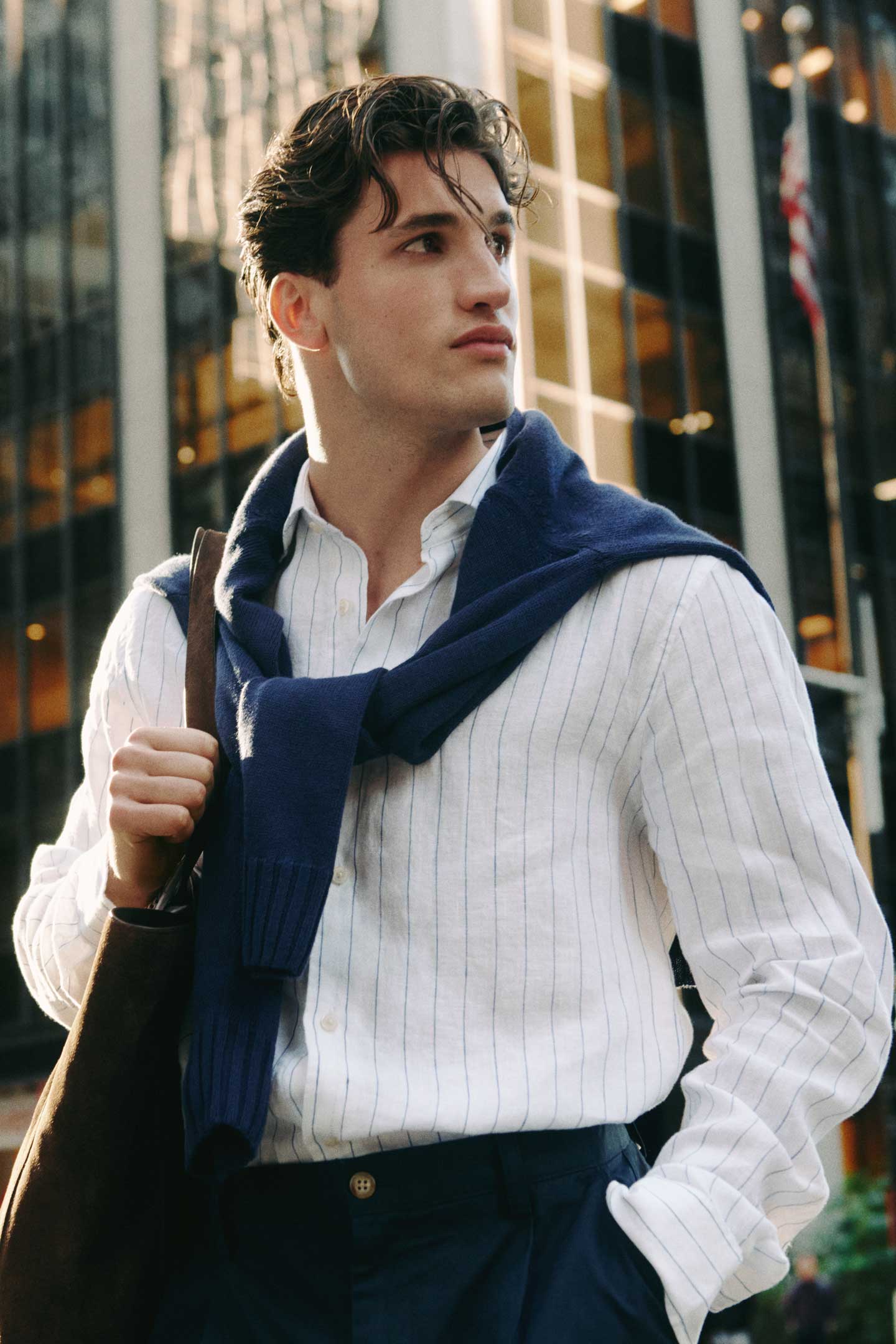 Man wearing a Gant white striped shirt with a navy sweater over the shoulders, holding a bag in the city.