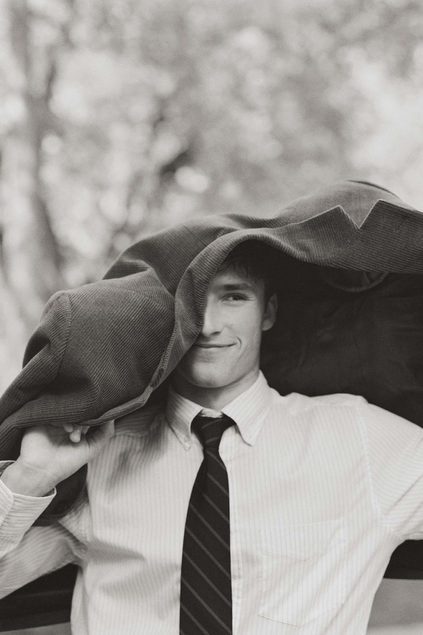 Black and white image of model styled in a GANT shirt with tie holding a blazer over head in an outdoor setting.