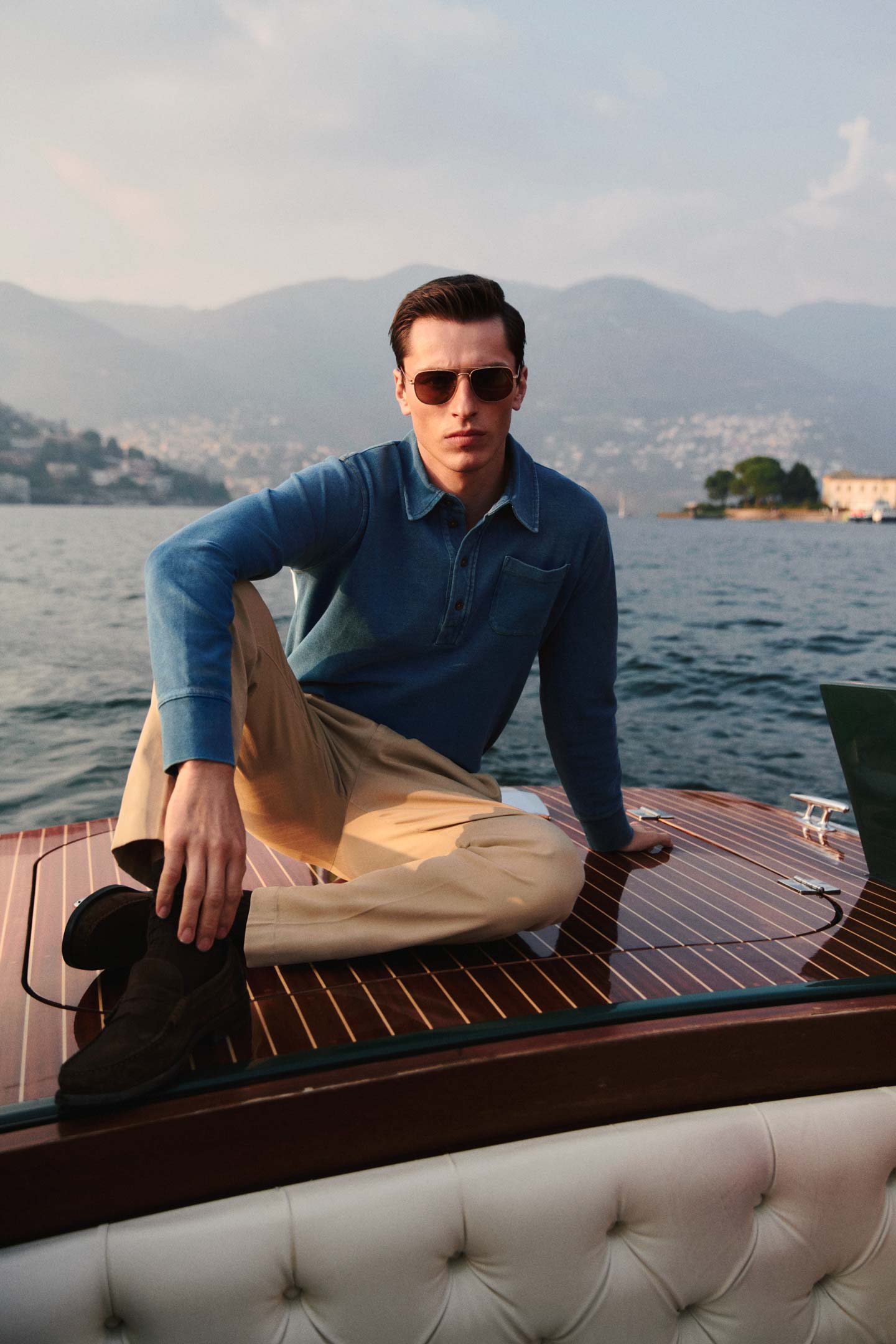 Model styled in a GANT blue rugger and beige trousers sitting on a wooden boat by the water.