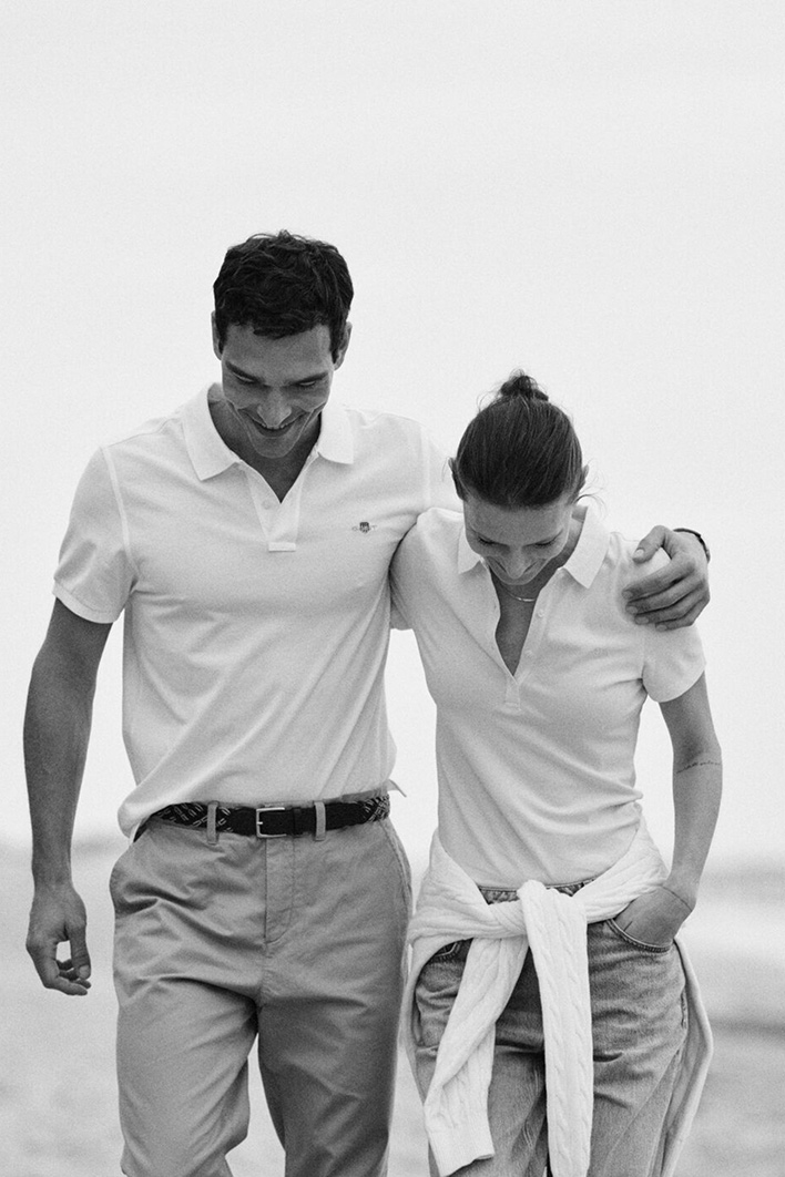 Two people wearing Gant white polo shirts walking arm in arm on a beach.