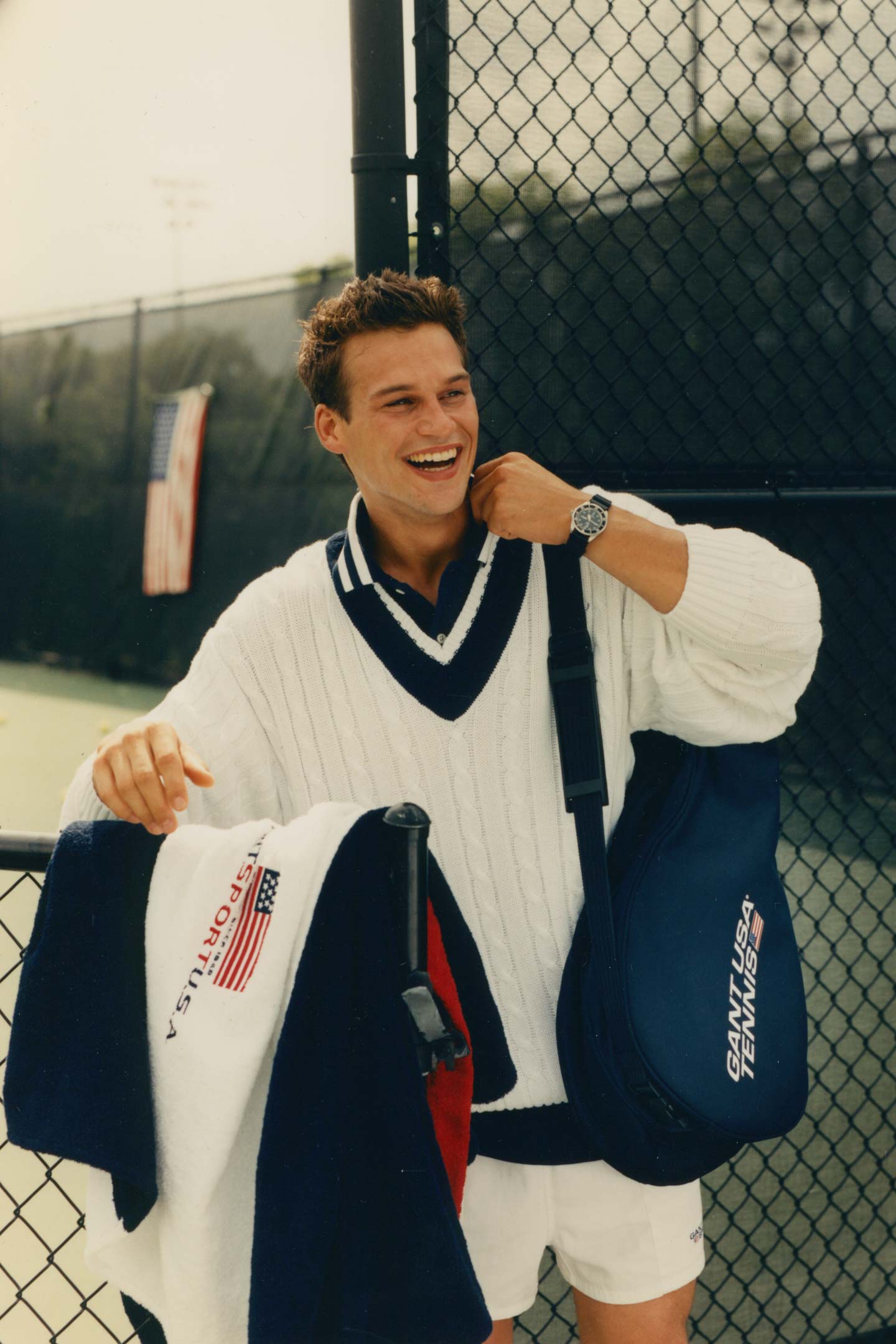 Man wearing a Gant tennis sweater and holding gear while smiling beside a court fence.