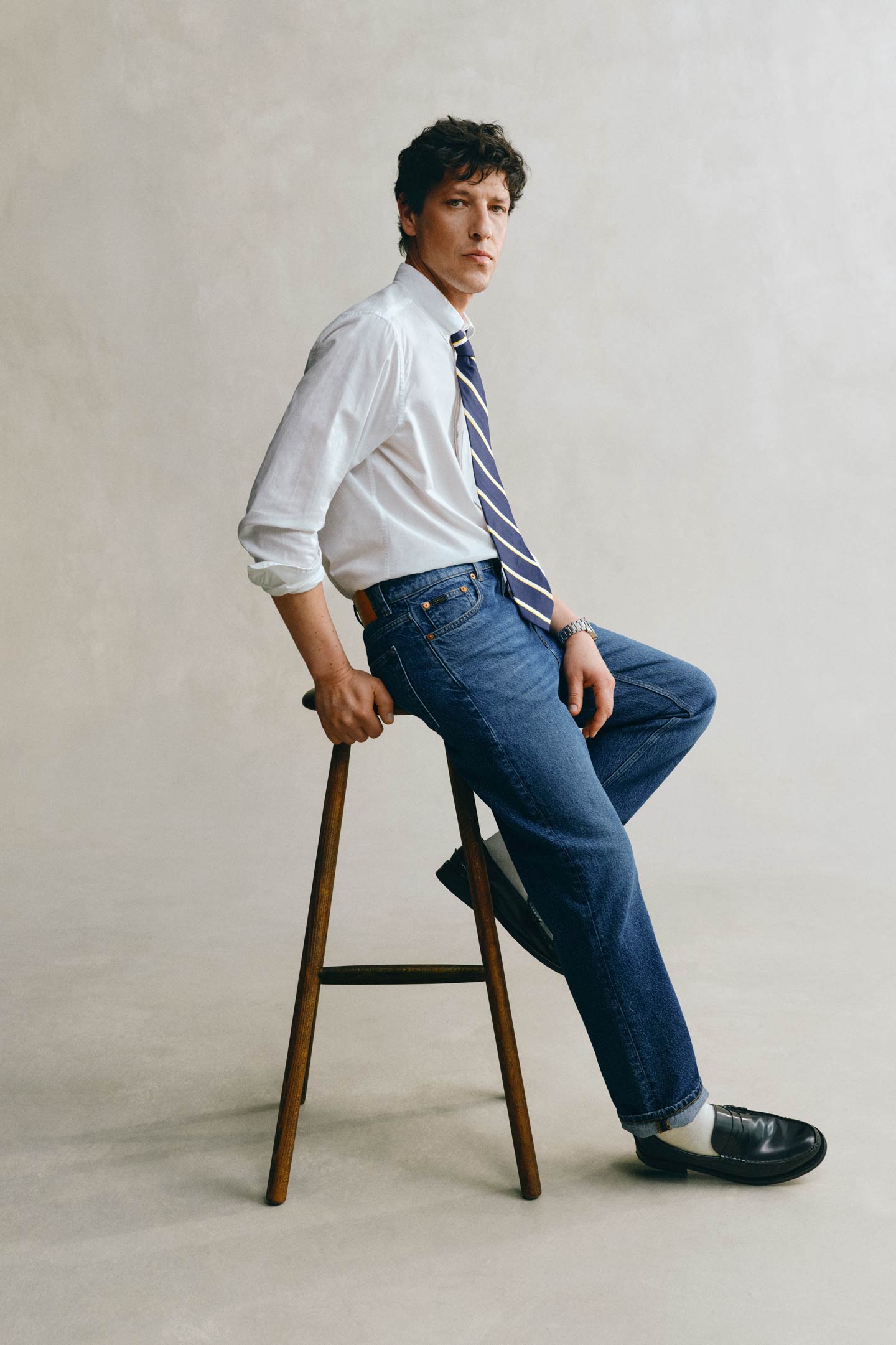 Model styled in a GANT white shirt, tie and blue jeans seated on a stool in front of a grey backdrop.