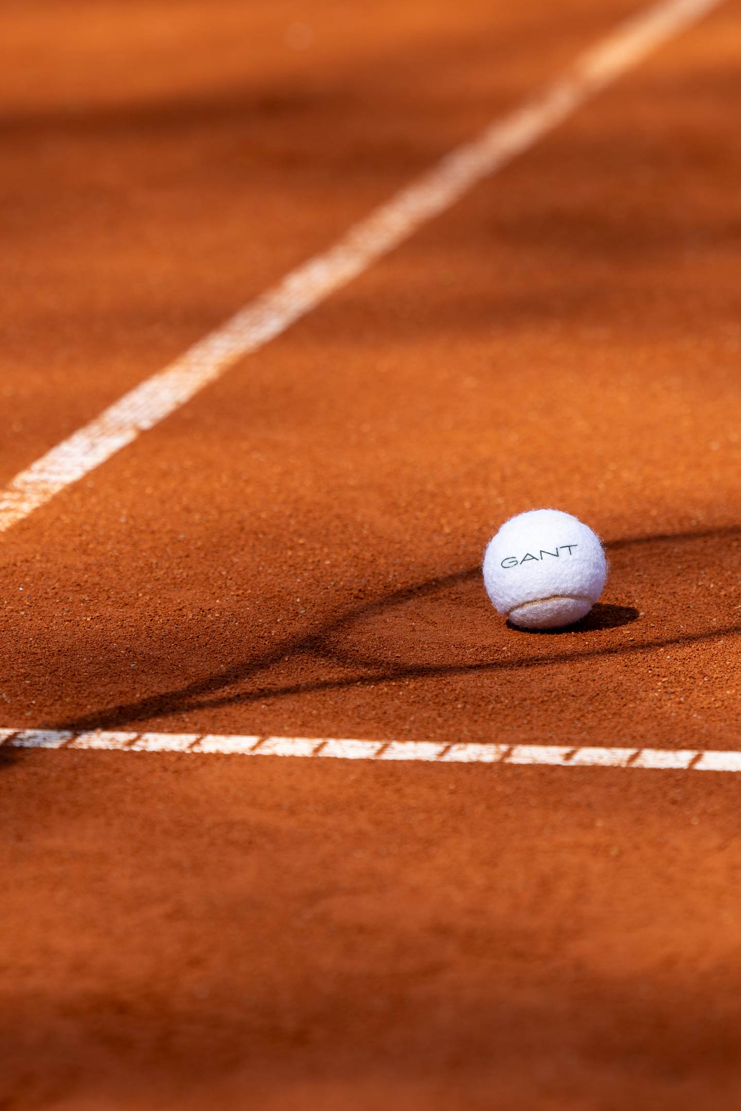 GANT tennis ball resting on a clay tennis court near the baseline.