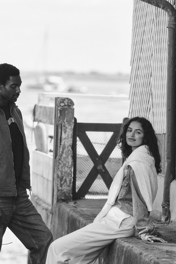 Due persone che parlano vicino a un edificio sul lungomare, indossando outfit casual a strati di Gant.