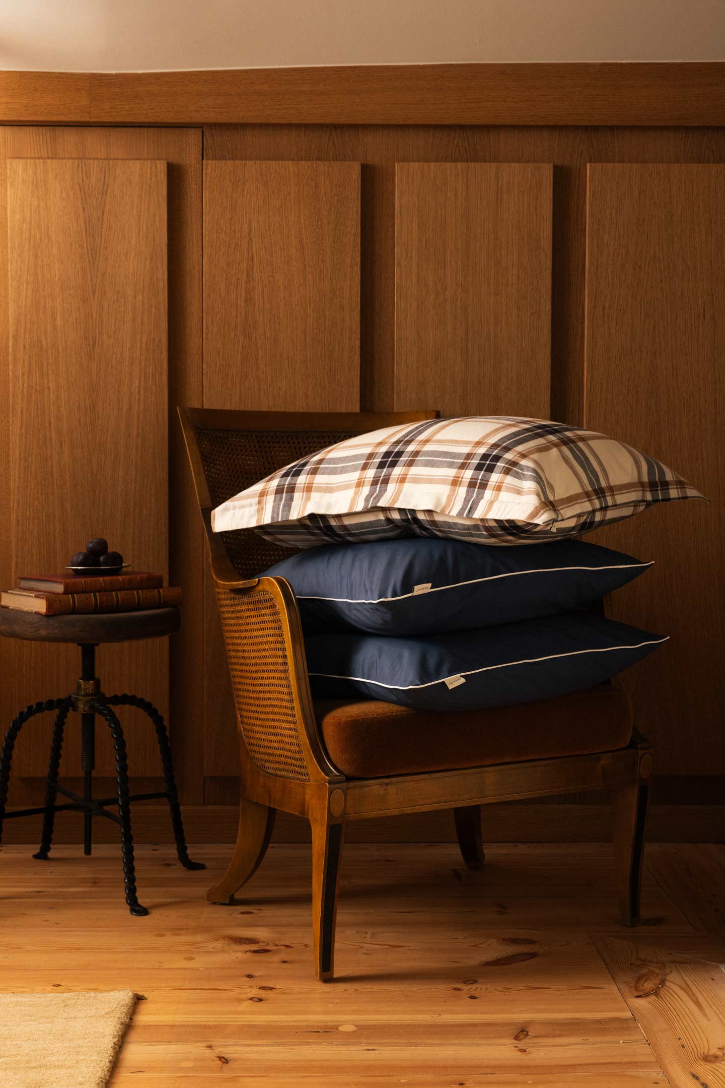 Gant checked and solid-colour pillows stacked on a wooden chair in a bedroom setting.
