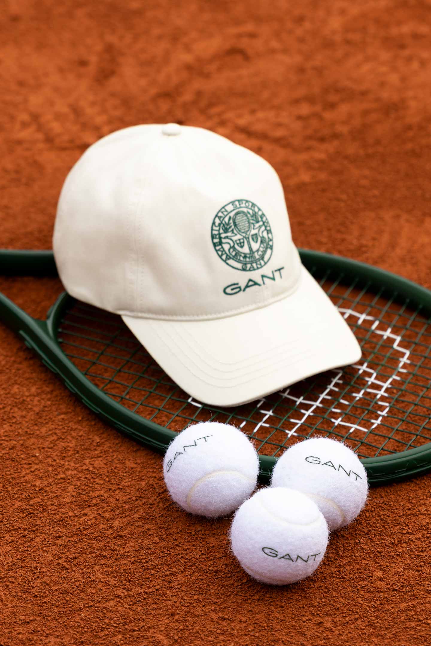 White Gant cap displayed on a tennis racket with branded tennis balls on a clay court.