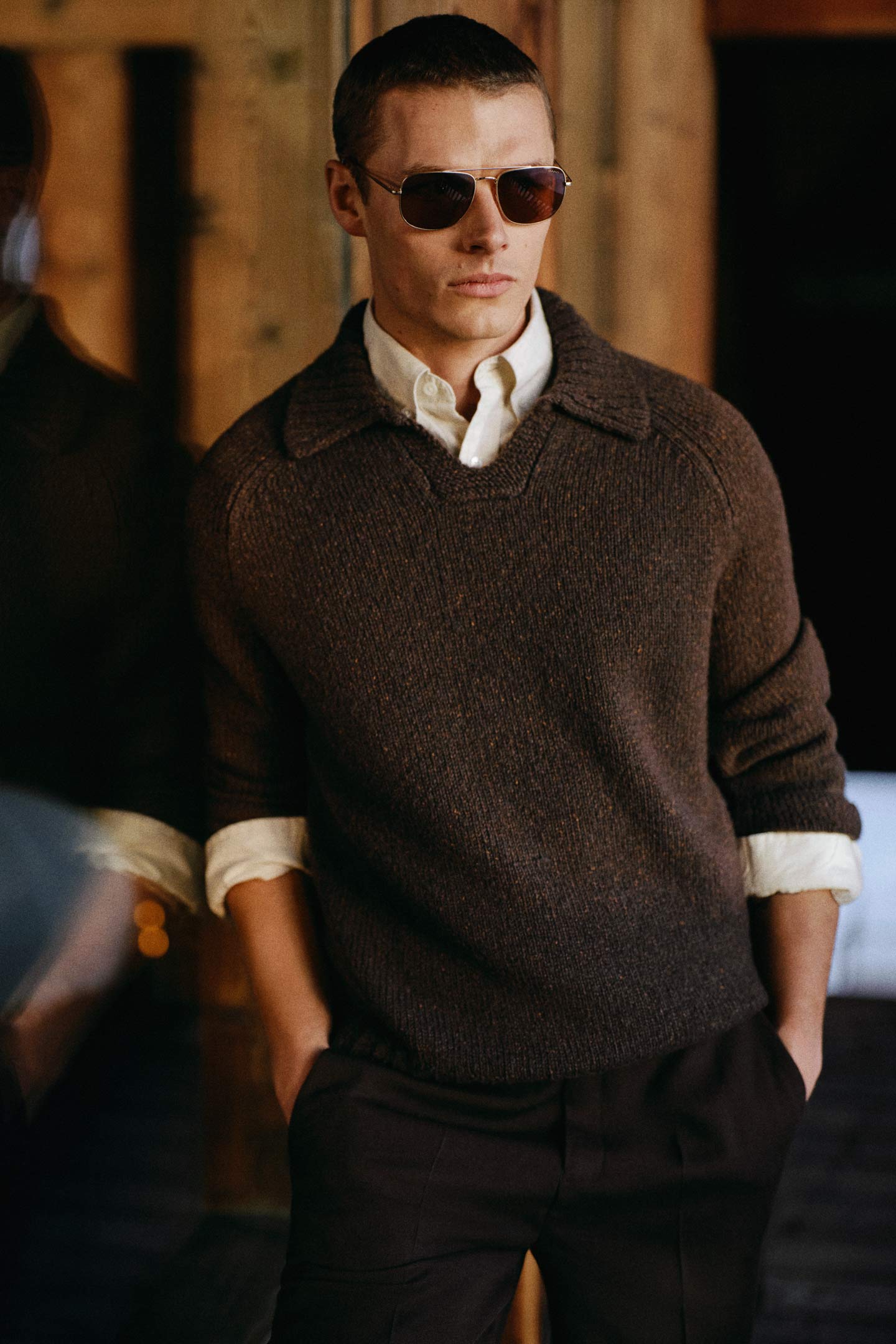 Model styled in a Gant collared knit sweater layered over a button-down shirt with tailored pants and sunglasses.