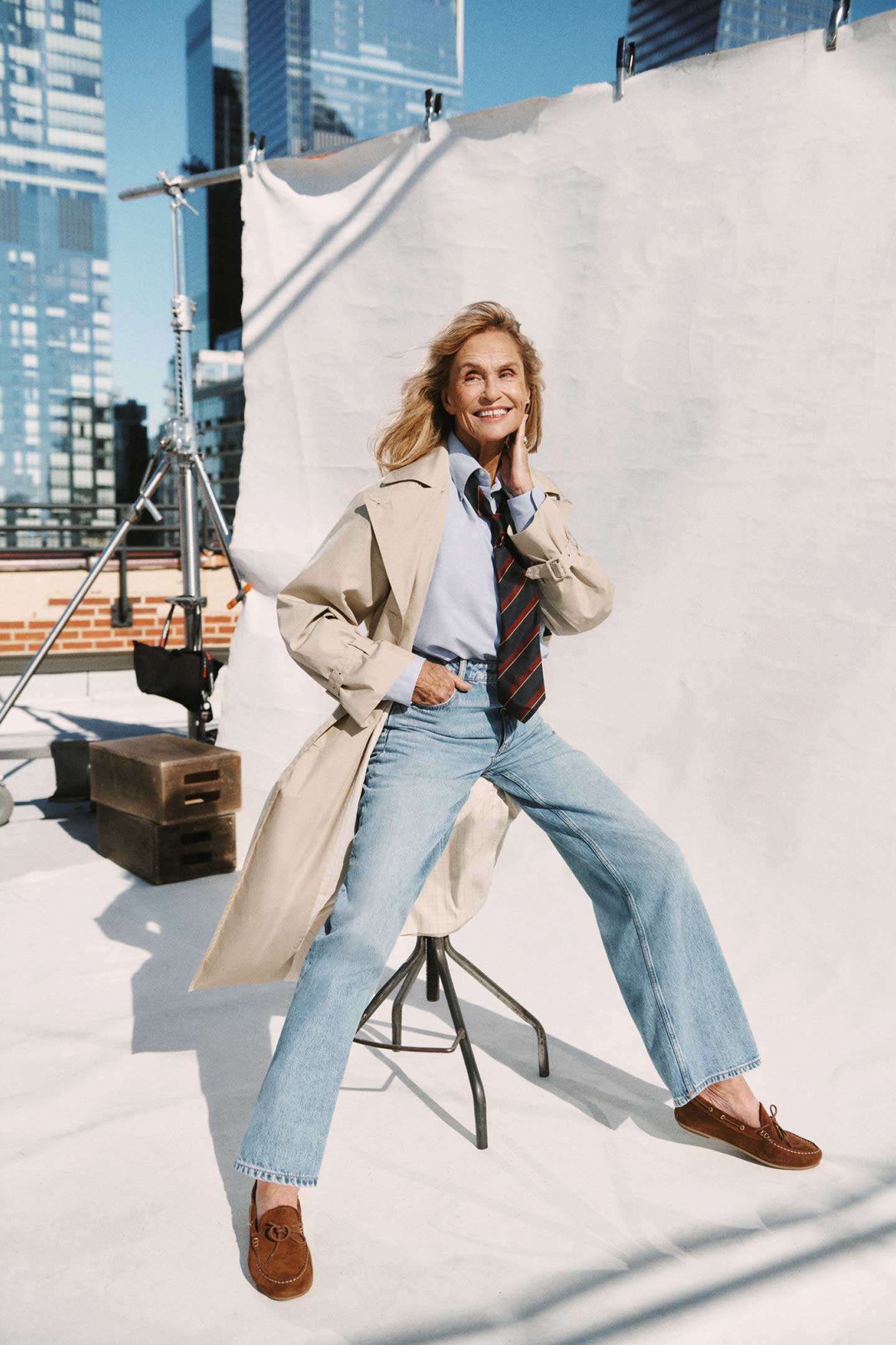 Woman wearing a Gant trench coat, shirt, tie, and jeans posing on a rooftop set with city buildings in the background