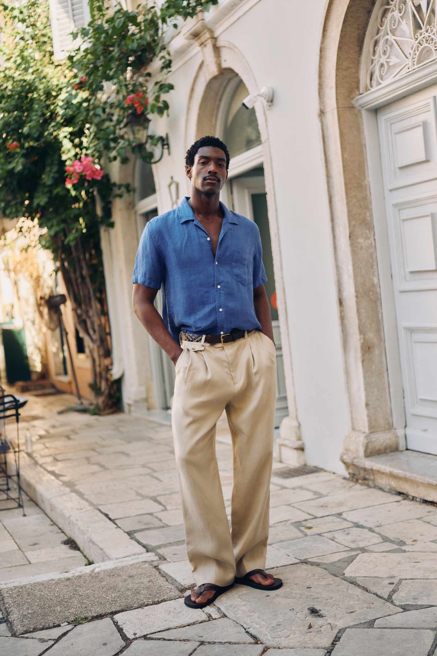 Man standing on a sunlit street wearing a short‑sleeve blue linen shirt paired with beige wide‑leg trousers and flip‑flops.