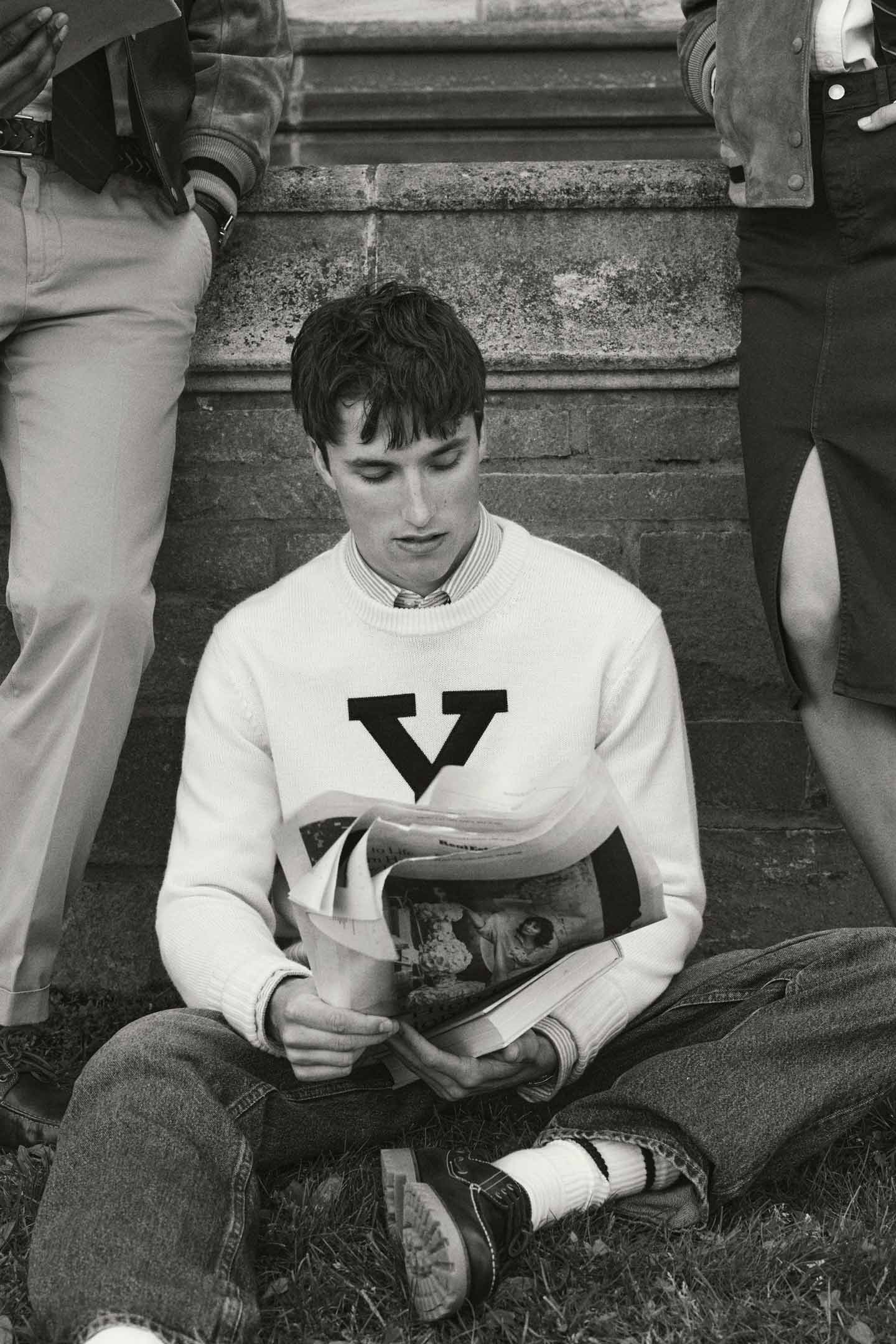 Black and white image of model styled in a GANT Yale sweatshirt, sitting on grass resting against a building.
