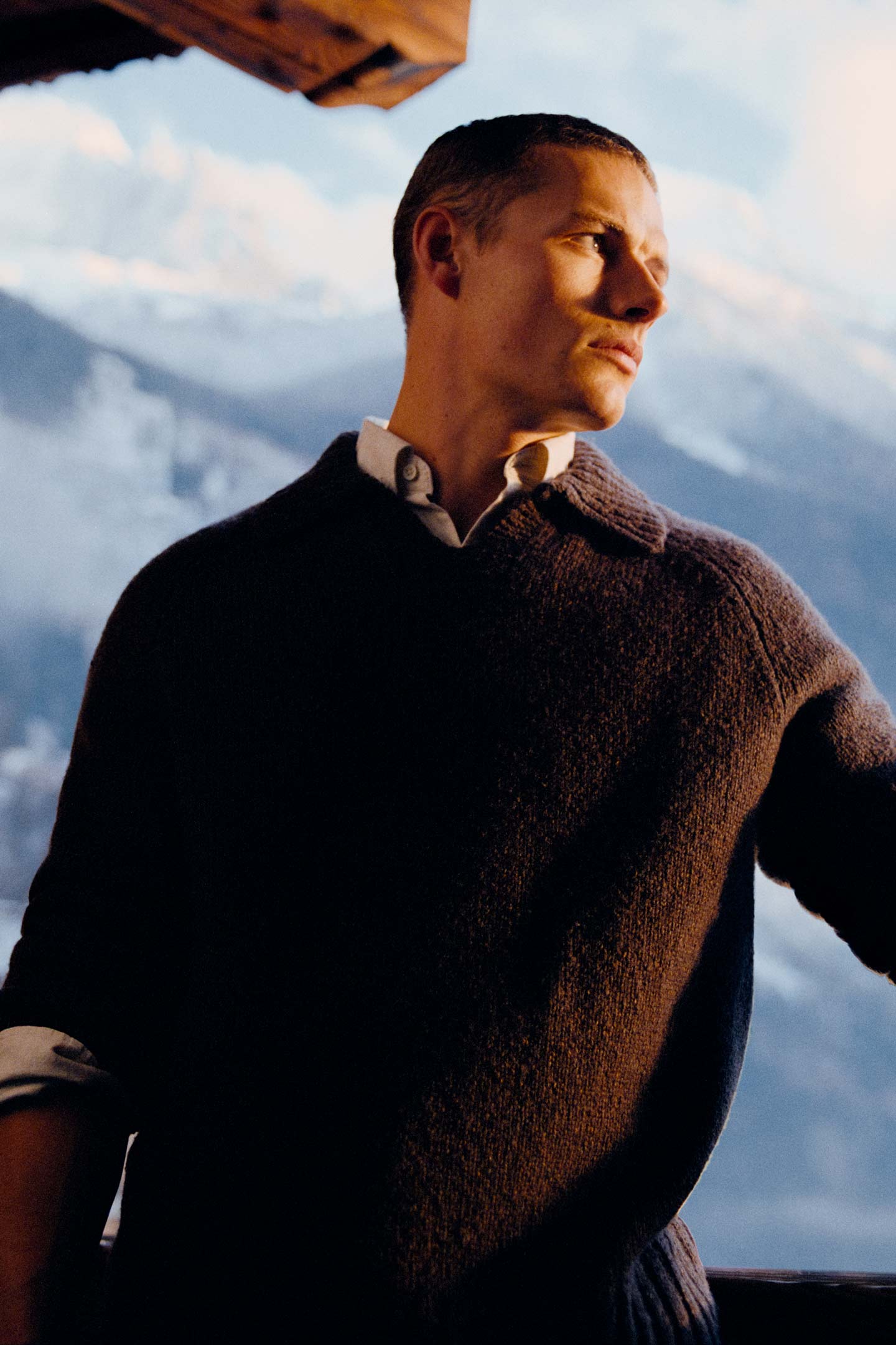 Model styled in a Gant collared knit sweater layered over a button-down shirt standing infront of a snowy mountain setting.