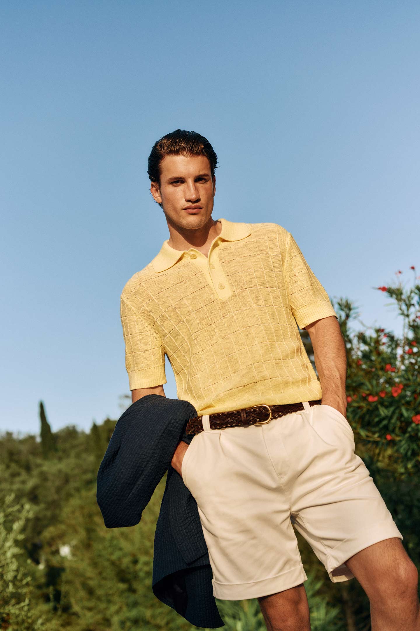 Men wearing a Gant yellow knit polo shirt with beige shorts and holding a dark jacket in an outdoor garden setting.