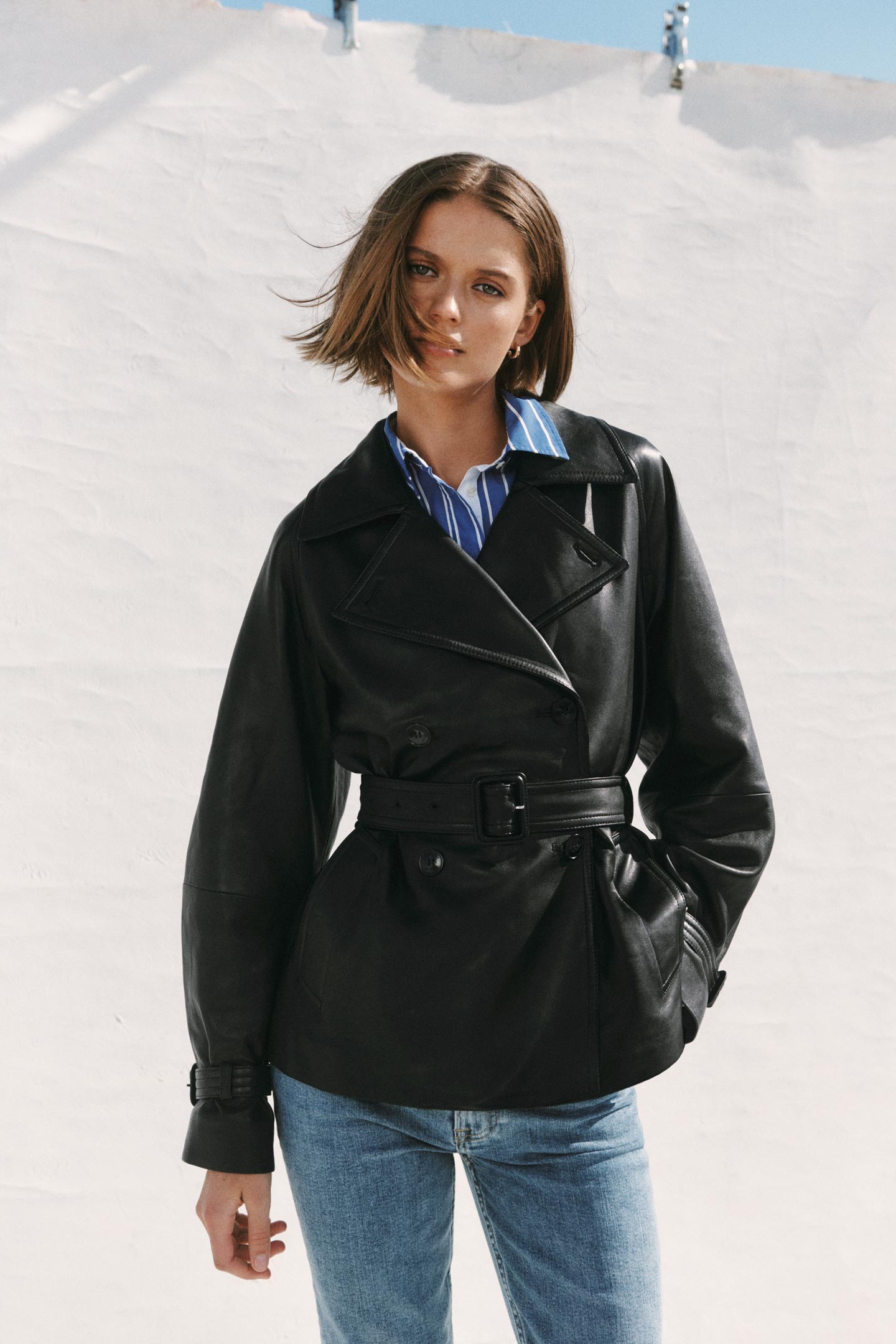 Person wearing a Gant black belted leather jacket with a striped shirt and jeans outdoors.