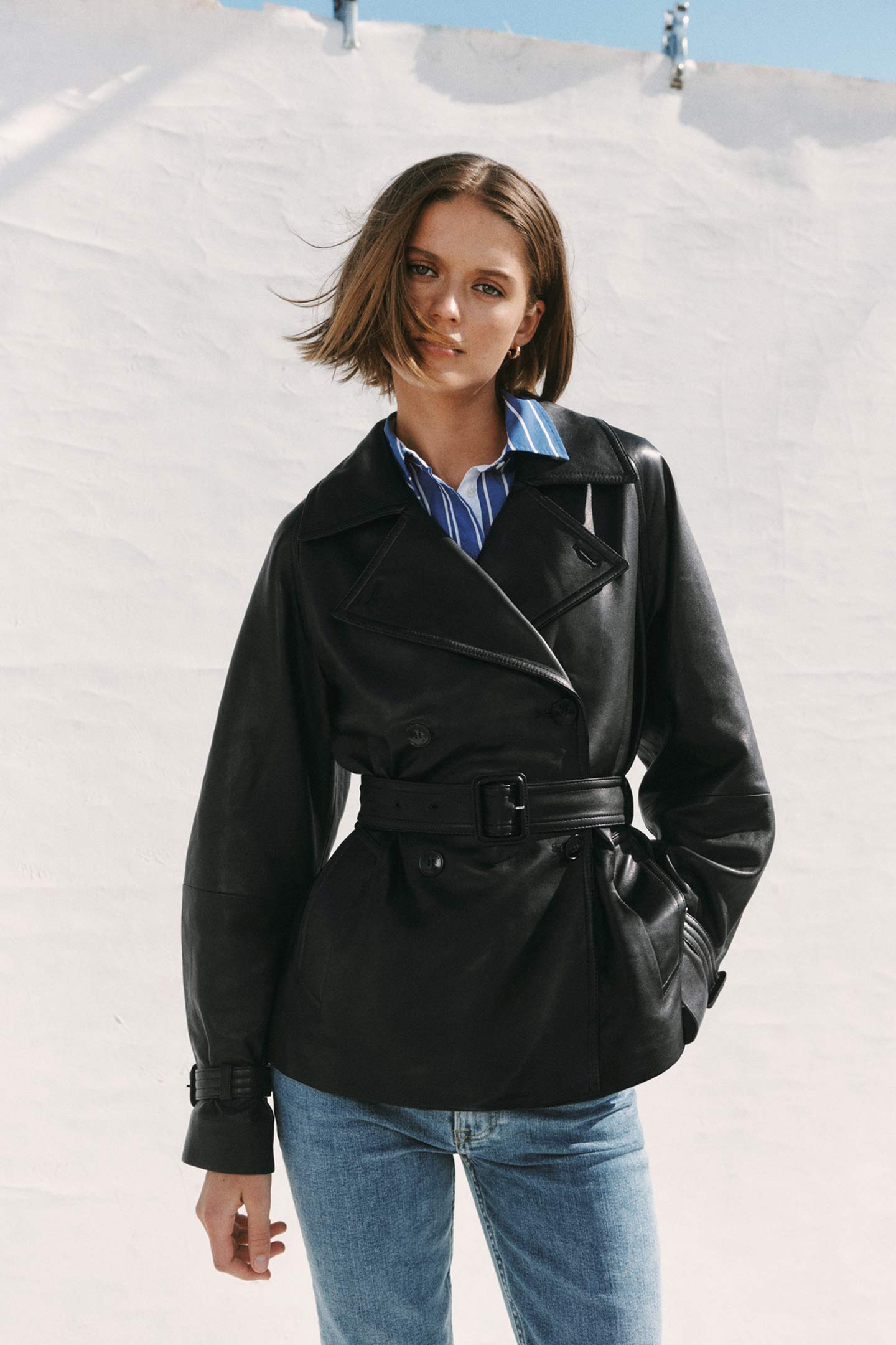 Persona con giacca di pelle nera Gant con cintura, camicia a righe e jeans all’aperto.