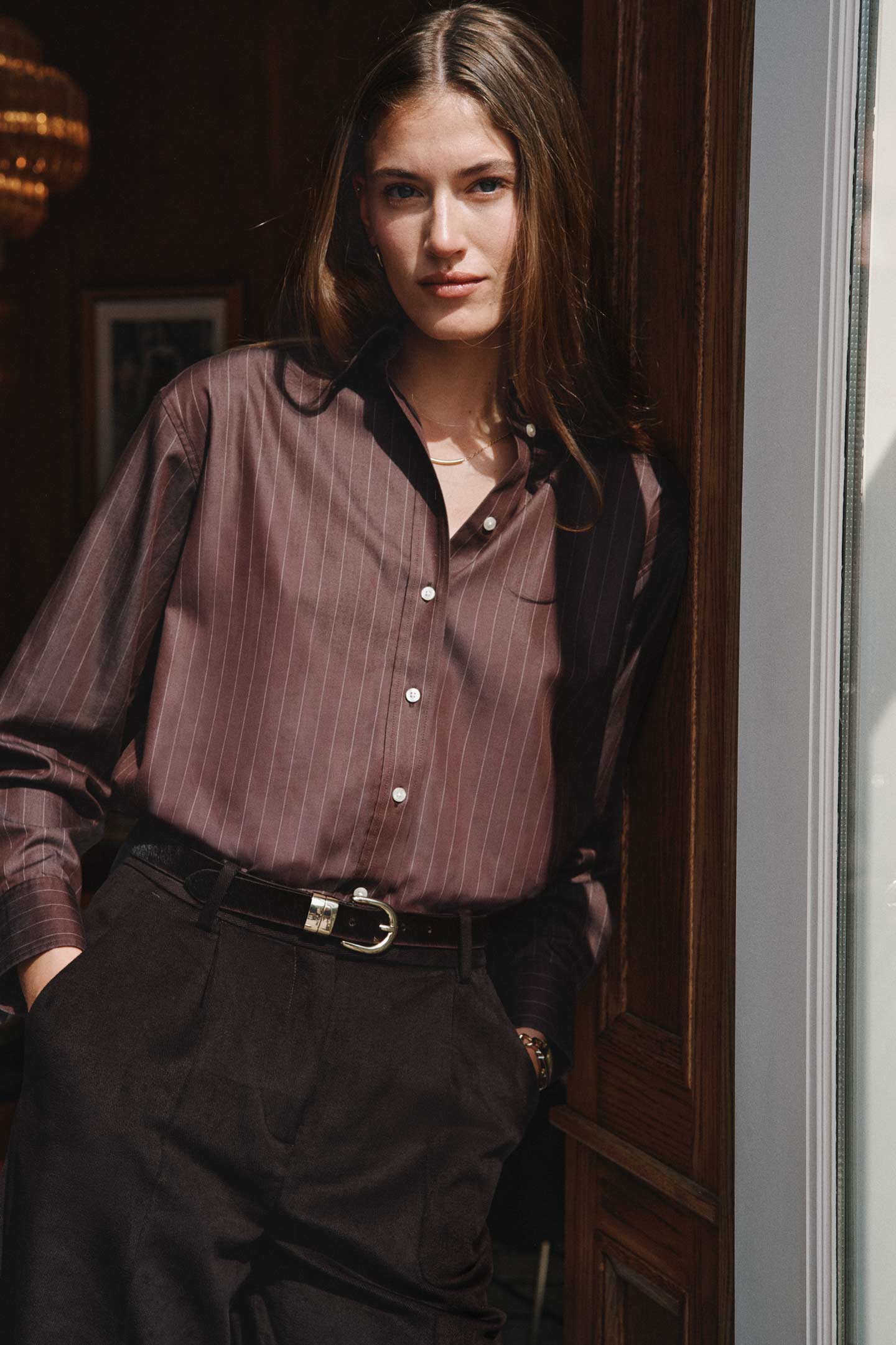 Model styled in a Gant striped button‑down shirt with dark tailored pants while standing in a doorway indoors.