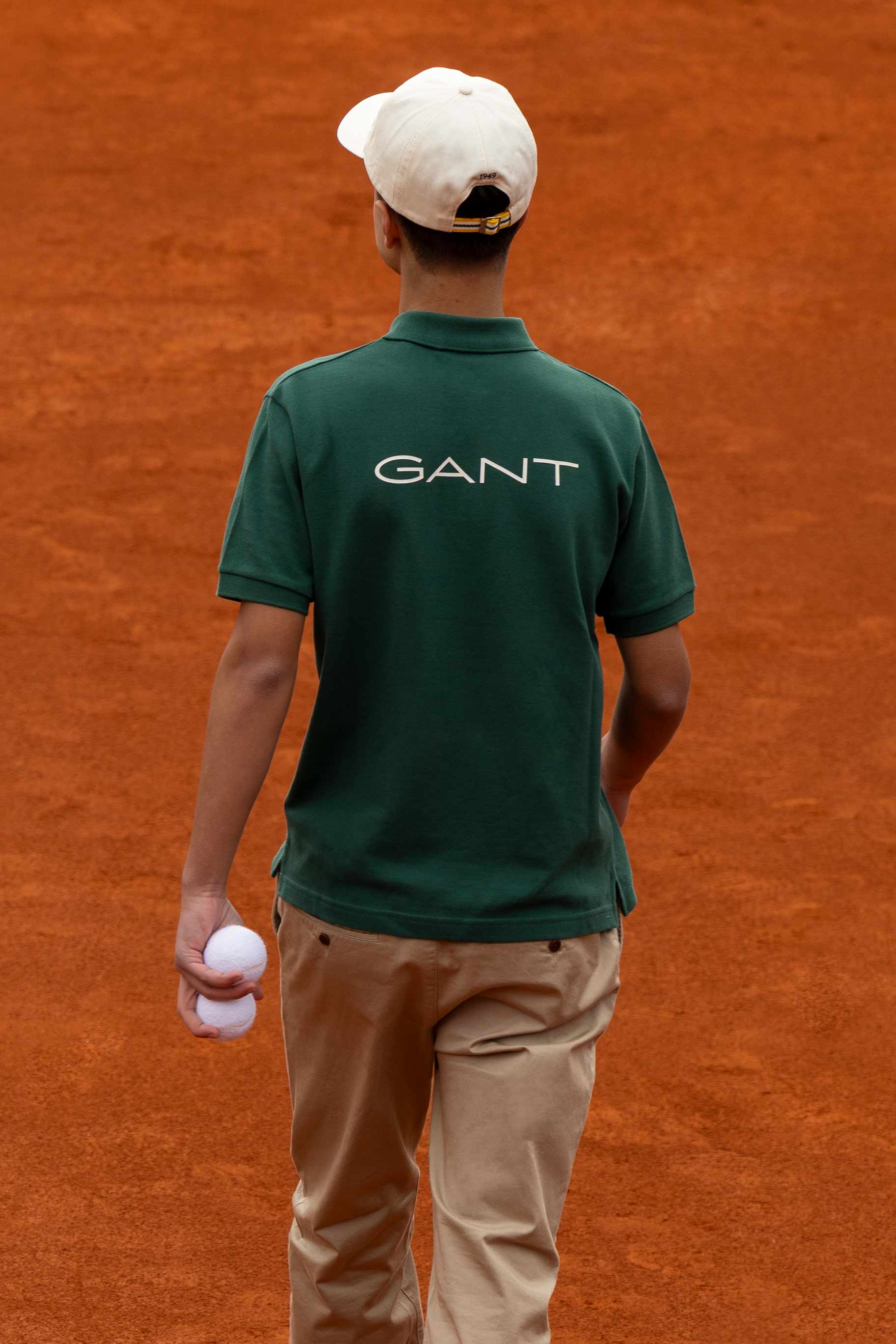 Two models wearing green GANT polo shirts with logo on the back, beige trousers, and white caps, standing on a clay tennis court.