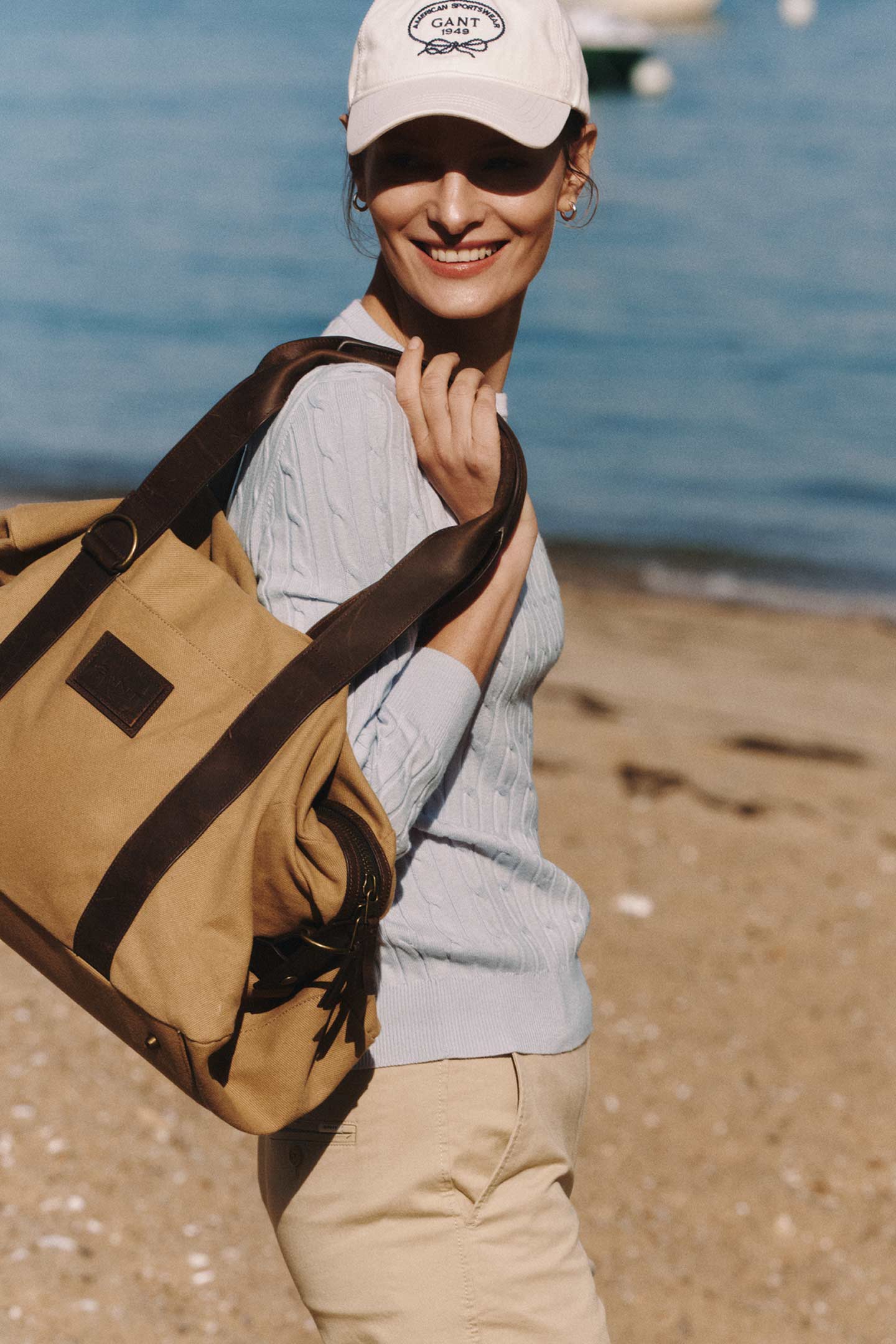 Woman wearing an Gant cable-knit sweater and cap, carrying a tan canvas shoulder bag on a beach.