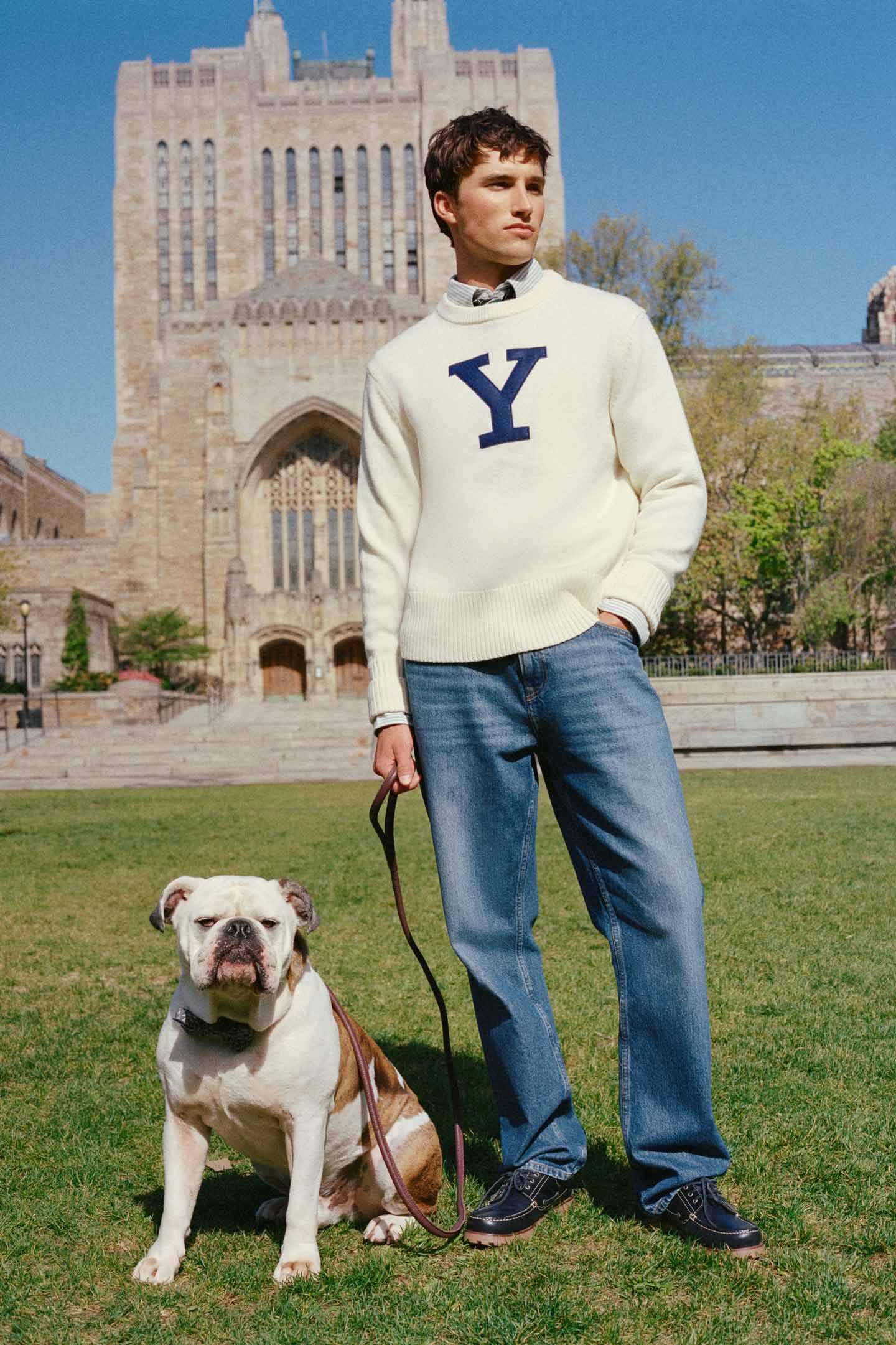 Model styled in a GANT Yale sweater and denim jeans standing with a dog in a campus setting.
