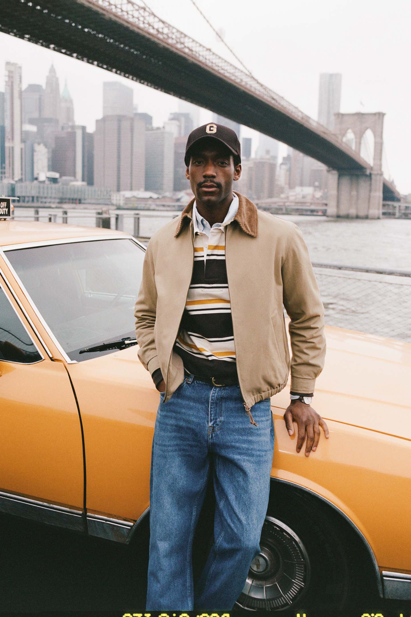Model styled in a Gant bomber jacket layered over a striped rugby shirt and jeans, standing beside a yellow taxi by the waterfront.