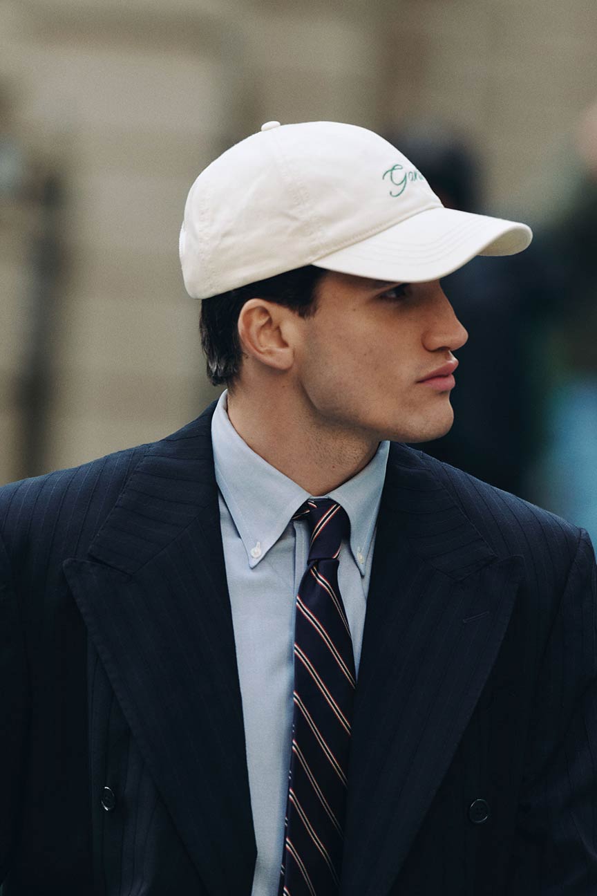 Man wearing a Gant pinstripe suit, collared shirt, striped tie and a white Gant cap, turning his head to the side outdoors