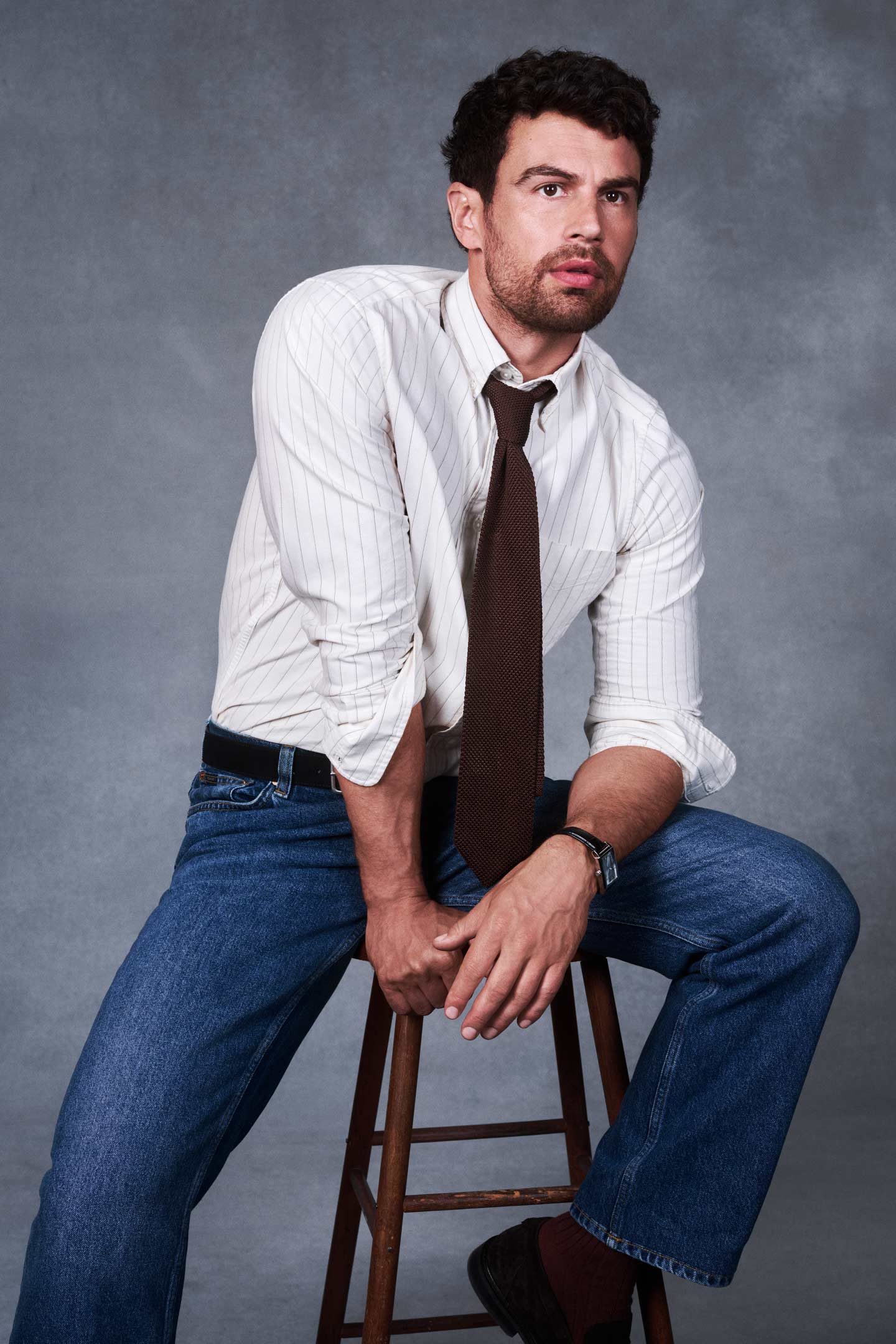 Actor Theo James styled in a Gant button-down shirt, tie and Gant jeans while seated on a stool in a studio setting.