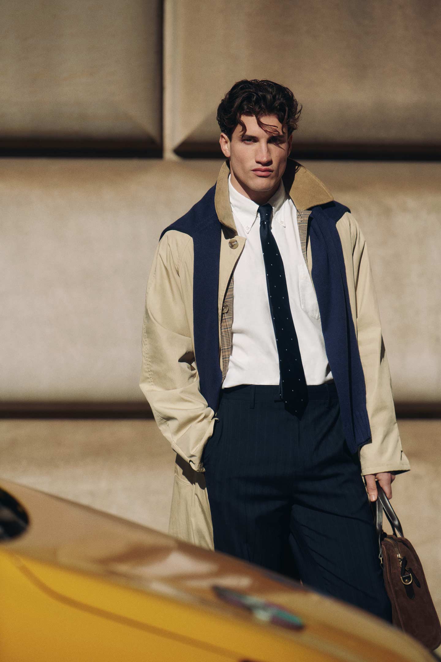 Personne portant un trench-coat kaki Gant sur une chemise blanche et une cravate, tenant un sac en cuir dans une rue de ville.