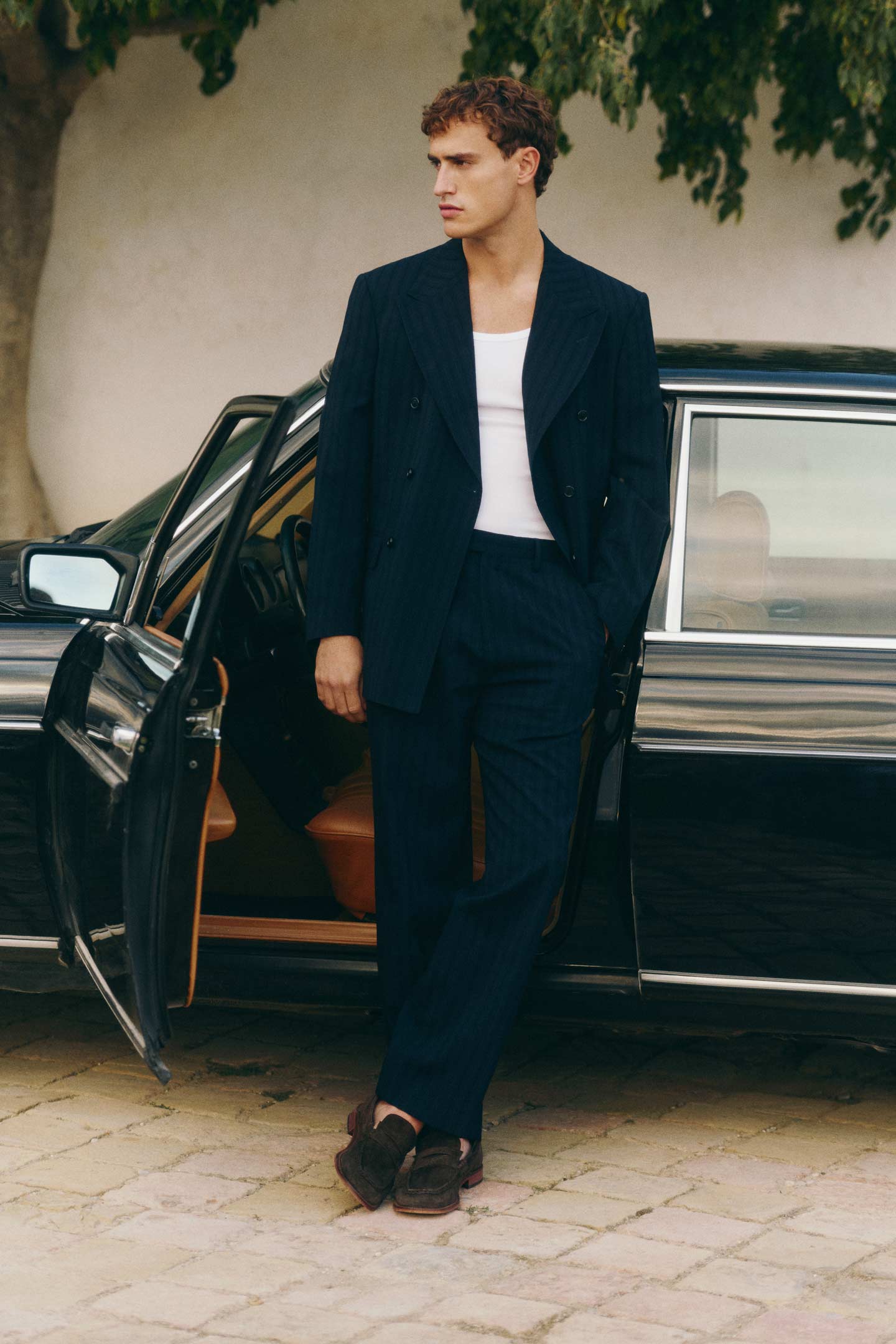 Man wearing a Gant navy suit and white tank top while standing beside an open car door.