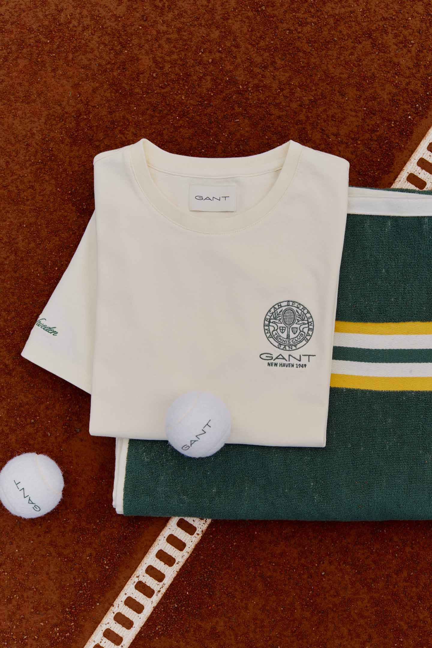 Gant T‑shirt and knit sweater with tennis balls displayed on a clay court beside a white line marker.