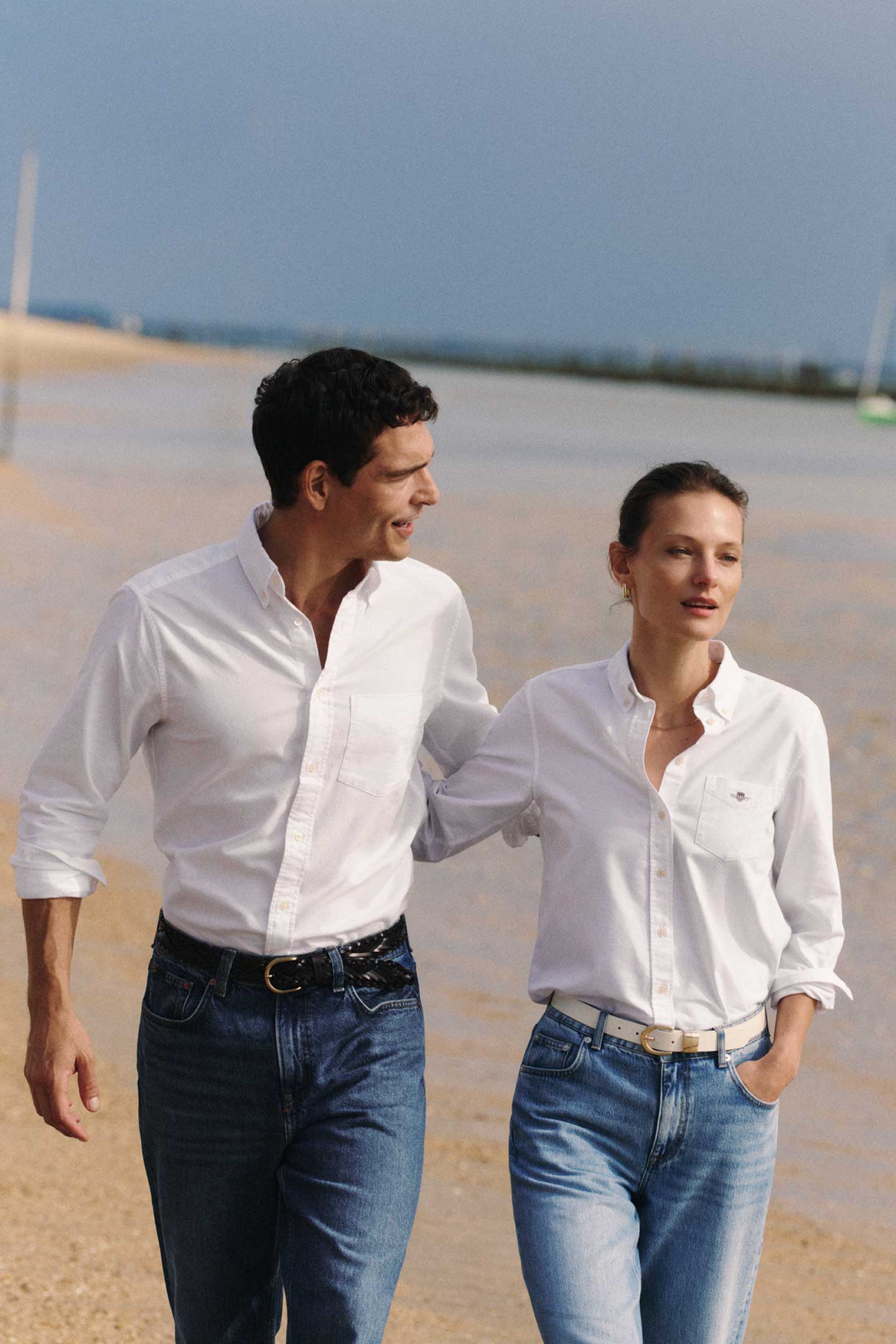 Deux personnes marchant sur la plage, portant des chemises blanches Gant et des jeans.
